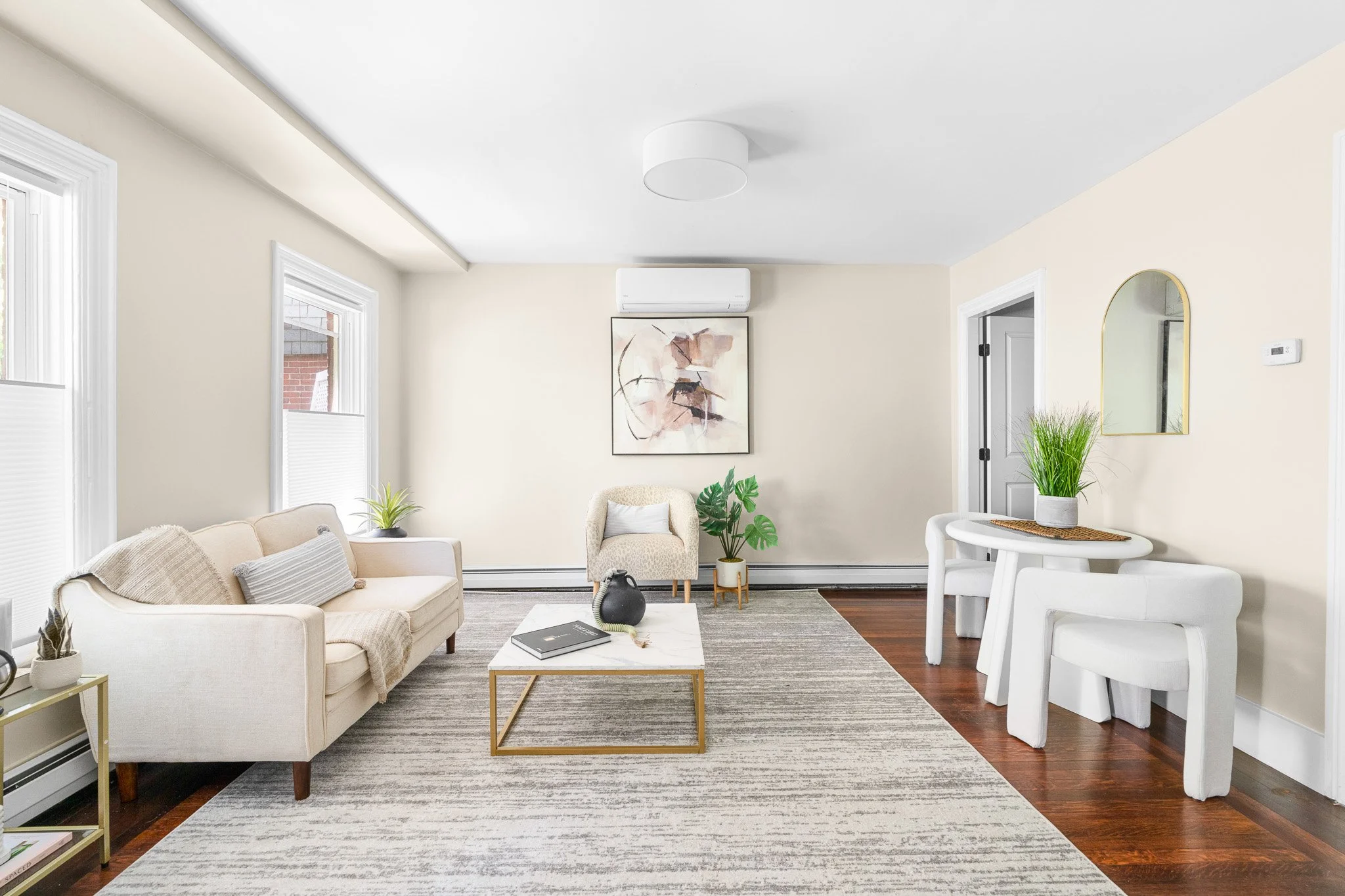 A living room with beige walls, white trim, and three windows with blinds. There is a beige sofa, an armchair, a white coffee table with a book and decorative object, a gray area rug, and multiple potted plants. Artwork hangs on the wall, and an air 