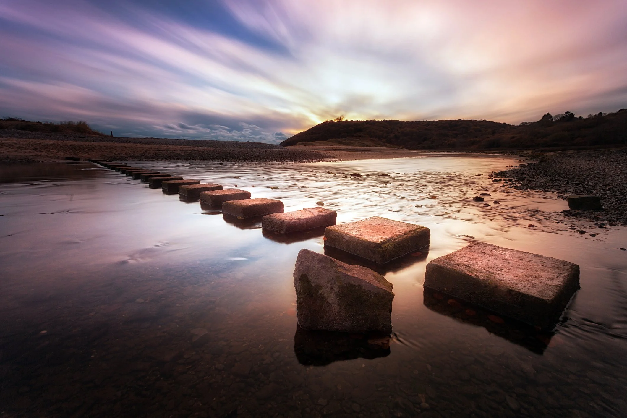 A peaceful river scene at sunset with a series of stepping stones leading across the water, surrounded by hills and a colorful sky with streaks of clouds.