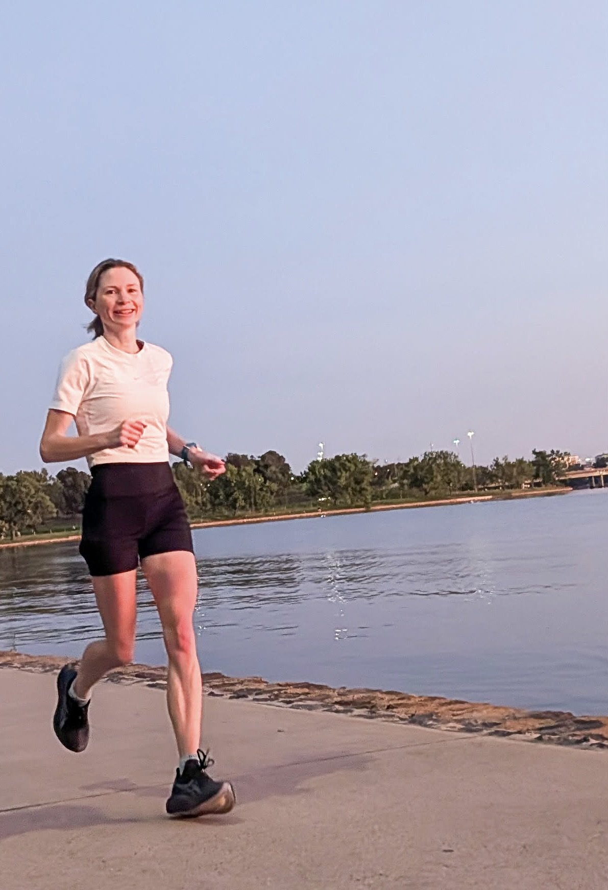 A woman jogging along a lakeside path during sunset, wearing a light pink t-shirt, black shorts, and running shoes.