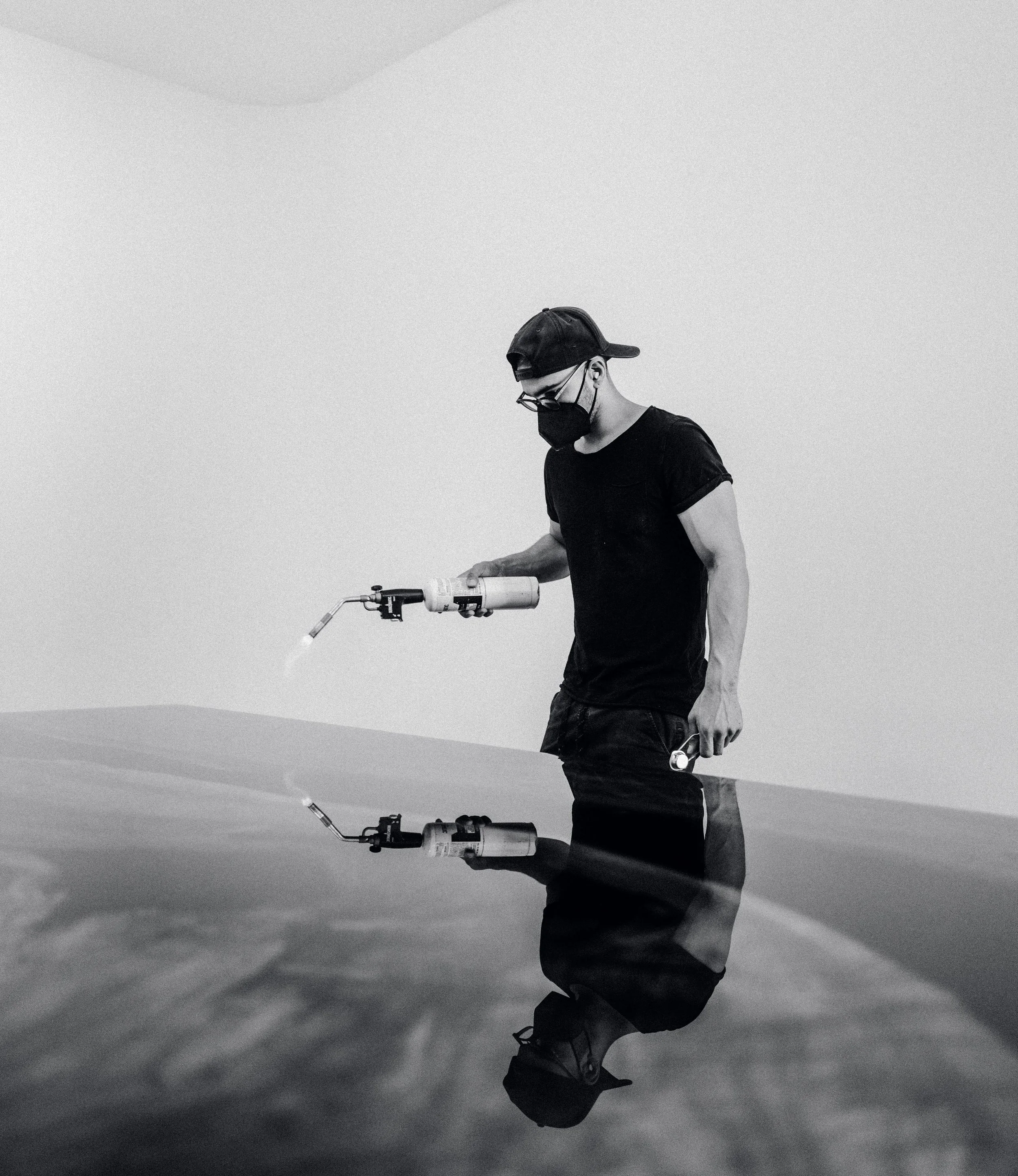 A person wearing a cap, glasses, and a face mask holding a blowtorch, seen as a reflection on a glossy surface.