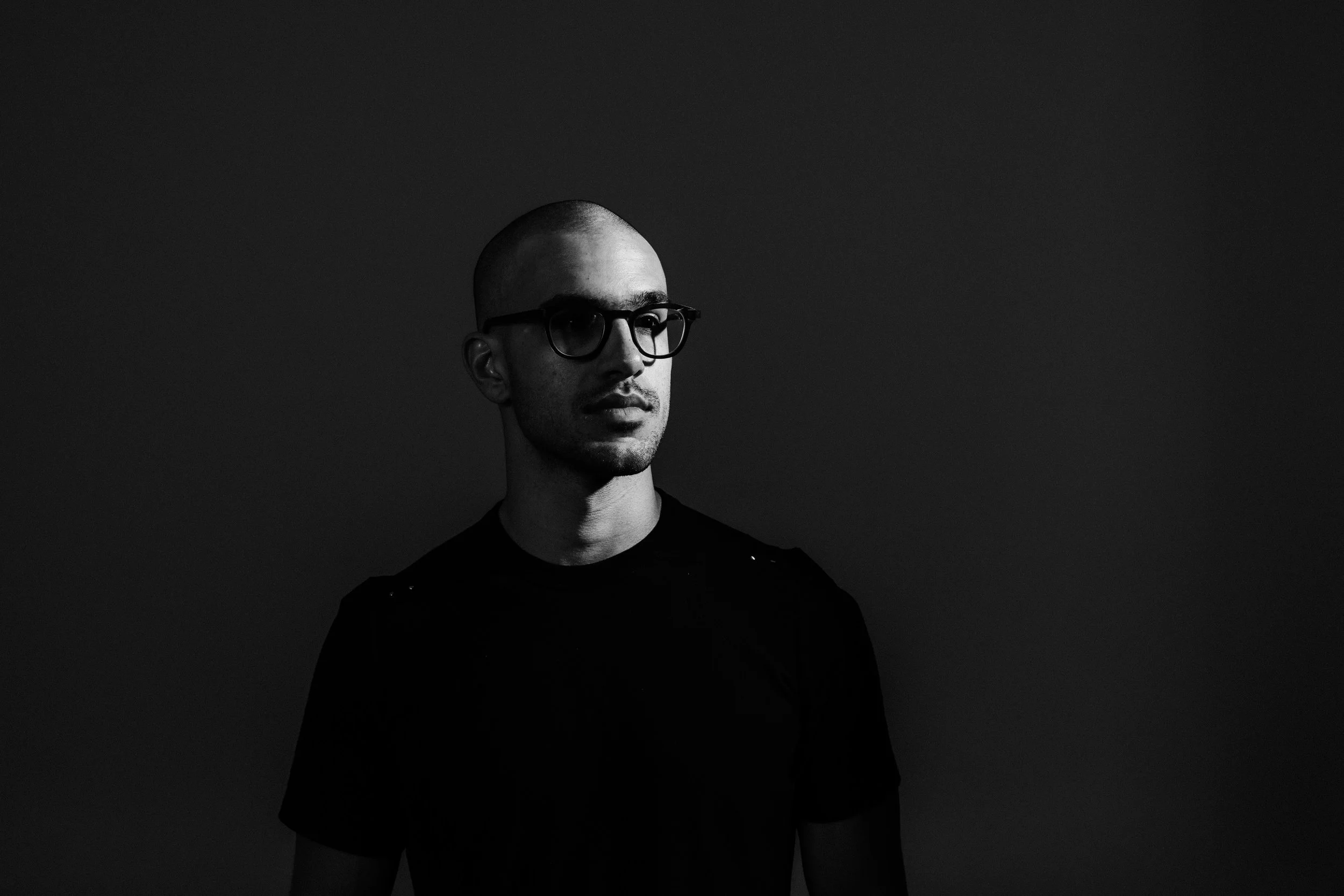 Black and white photo of a young man with a shaved head and glasses, looking to his right, against a dark background.