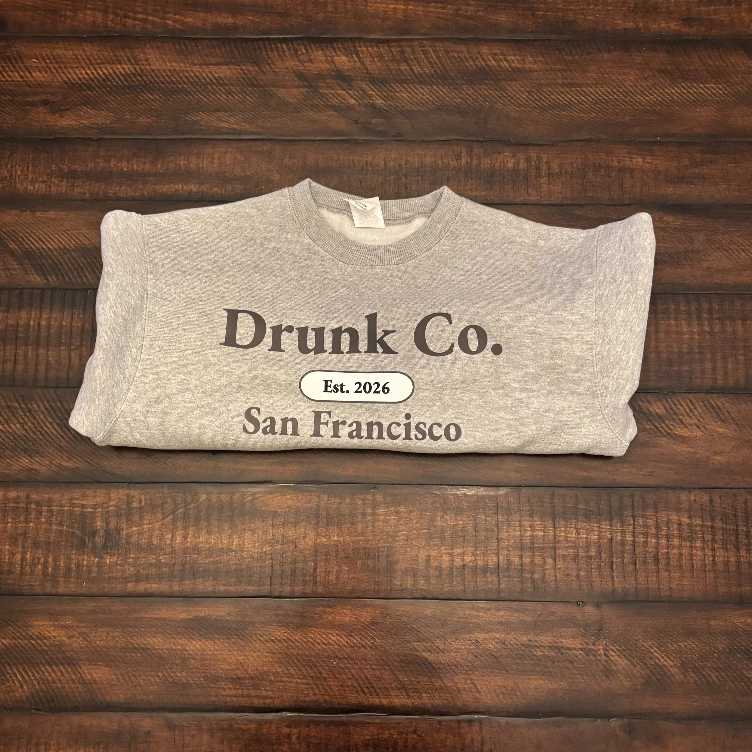 Gray T-shirt with black text that reads 'Drunk Co.' lying on a wooden surface.