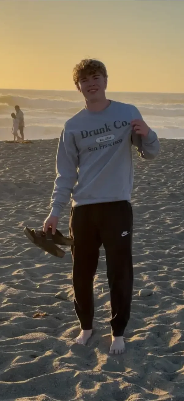 Teenager standing barefoot on sandy beach at sunset, holding sandals in one hand, smiling at the camera, with ocean waves in the background.