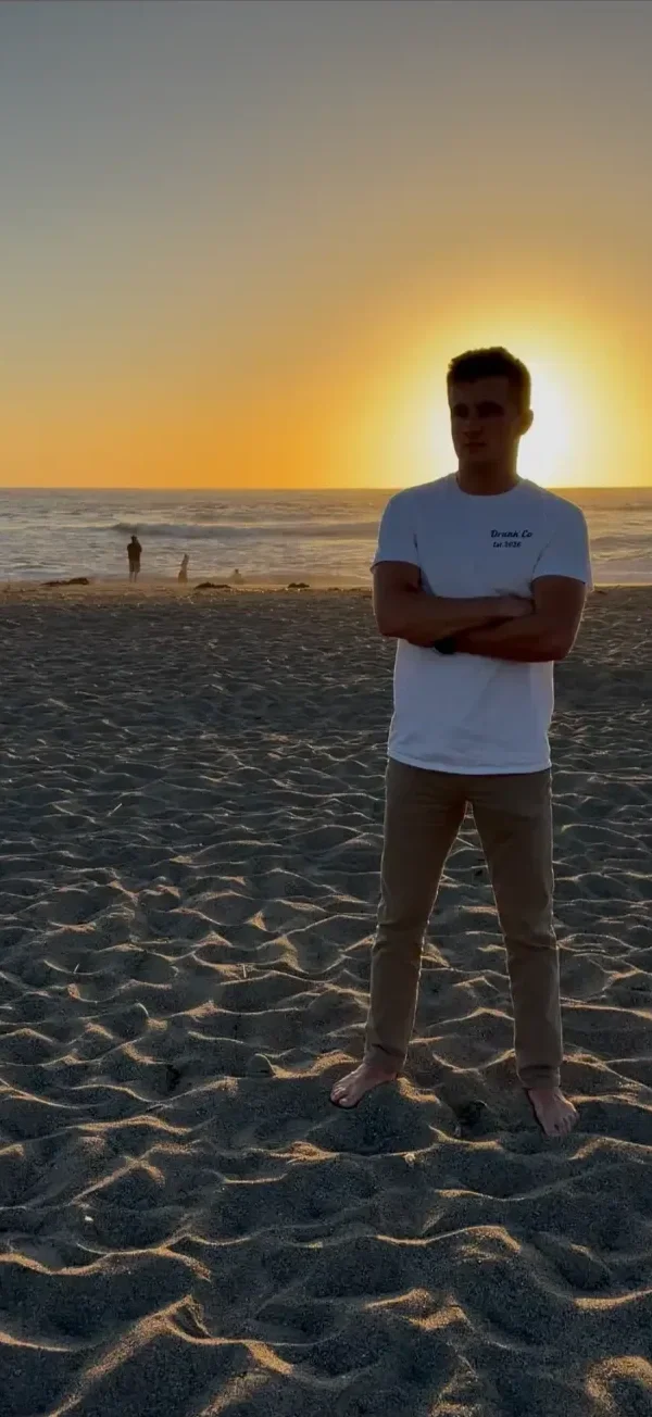 A young man stands barefoot on a sandy beach during sunset with the ocean in the background, arms crossed, facing the camera.