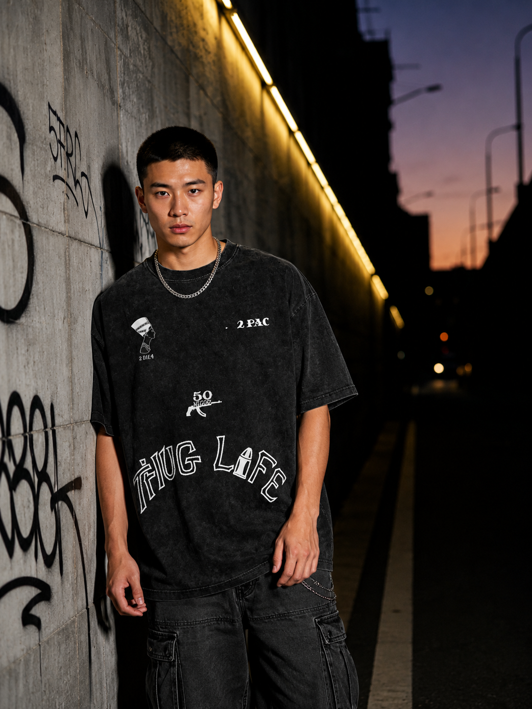 Young man with dark hair wearing black graphic T-shirt and black pants standing against a graffiti-covered wall during sunset.