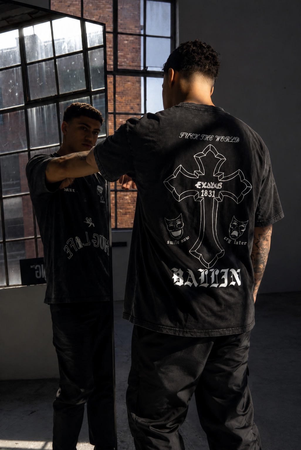 A person with short curly hair looking in a mirror, wearing a black graphic t-shirt and black pants, in front of industrial-style windows with brick and metal framing, with sunlight streaming in.