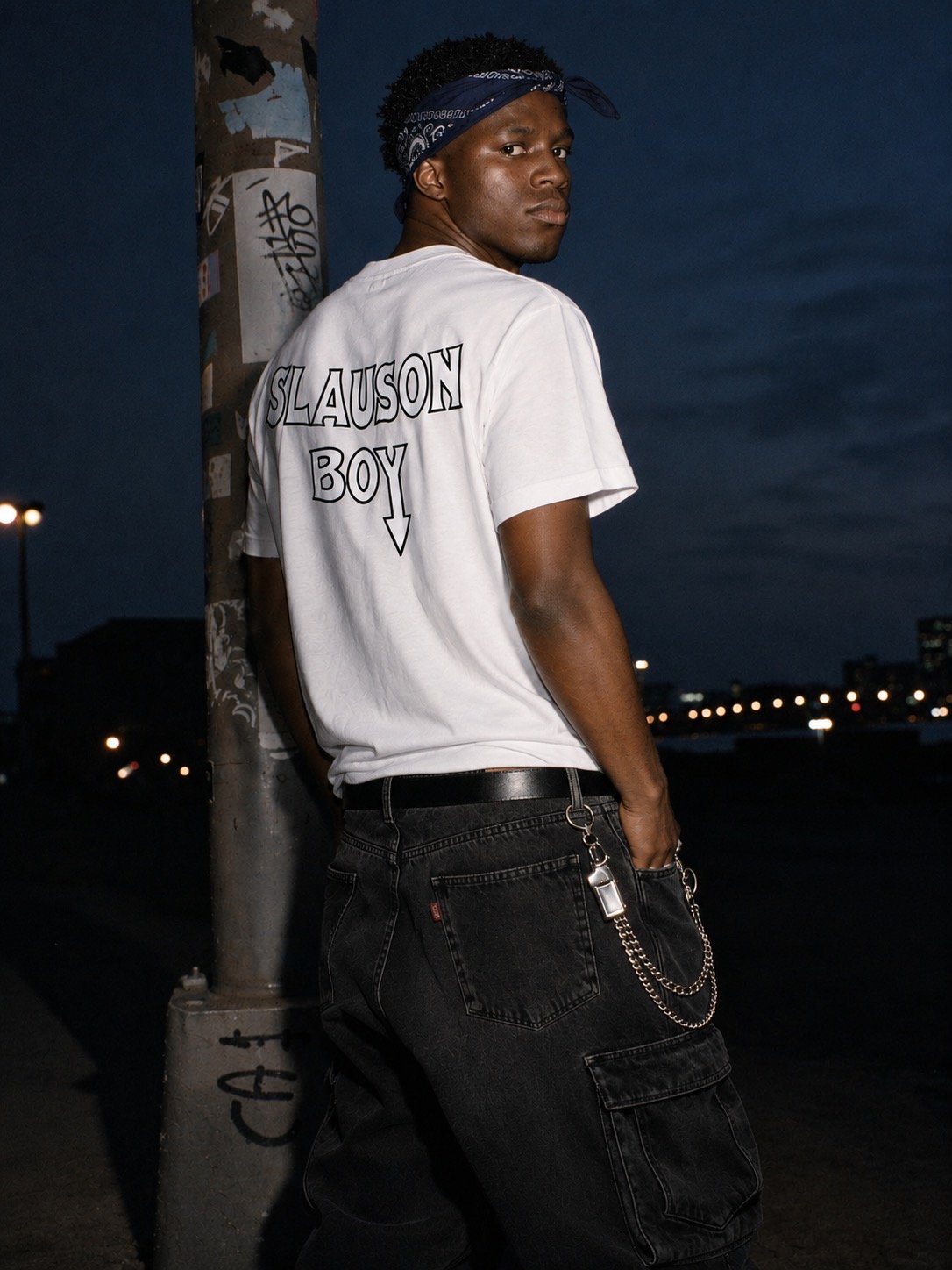 A young man stands next to a graffiti-covered lamppost, wearing a white T-shirt with 'SLAUSON BOY' written on the back and black pants with chains, against a dark nighttime cityscape background.