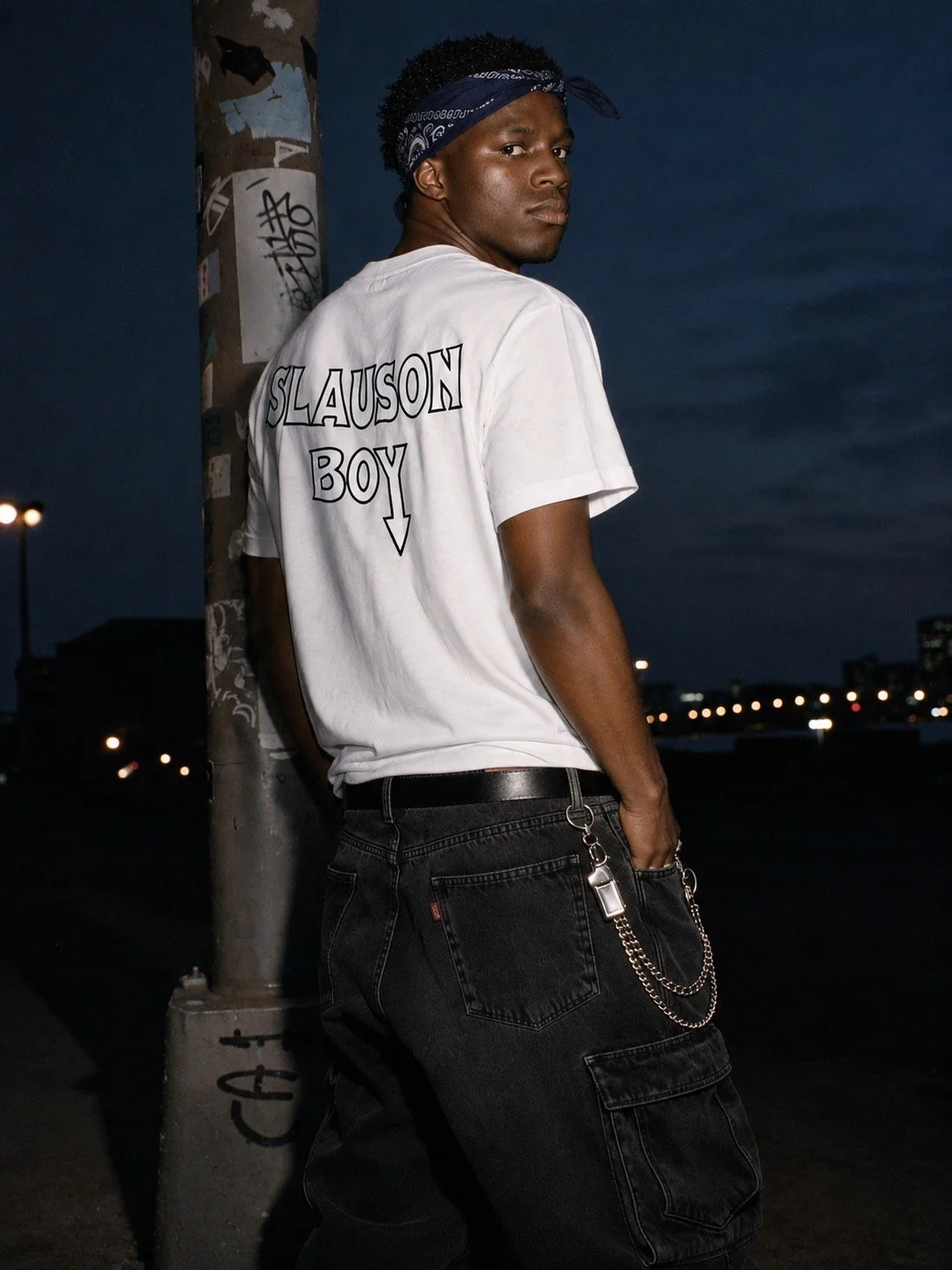 A young man with dark skin wearing a bandana, a white t-shirt with black text reading 'SLAUSON BOY' and a downward arrow, and black cargo pants with chains hanging from the pocket, standing near a graffiti-covered pole at night with city lights in the background.