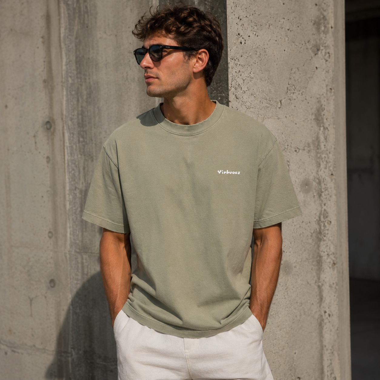 A young man in sunglasses and a green T-shirt with white text, standing against a concrete wall, hands in pockets.