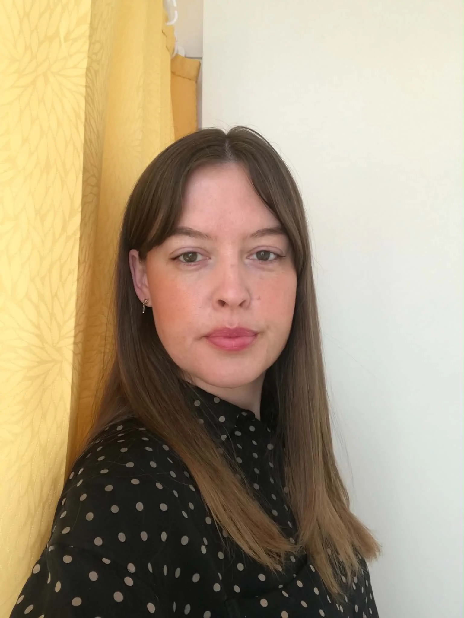 Portrait of a young woman with long brown hair, wearing a black polka dot shirt, standing indoors against a yellow curtain and white wall.