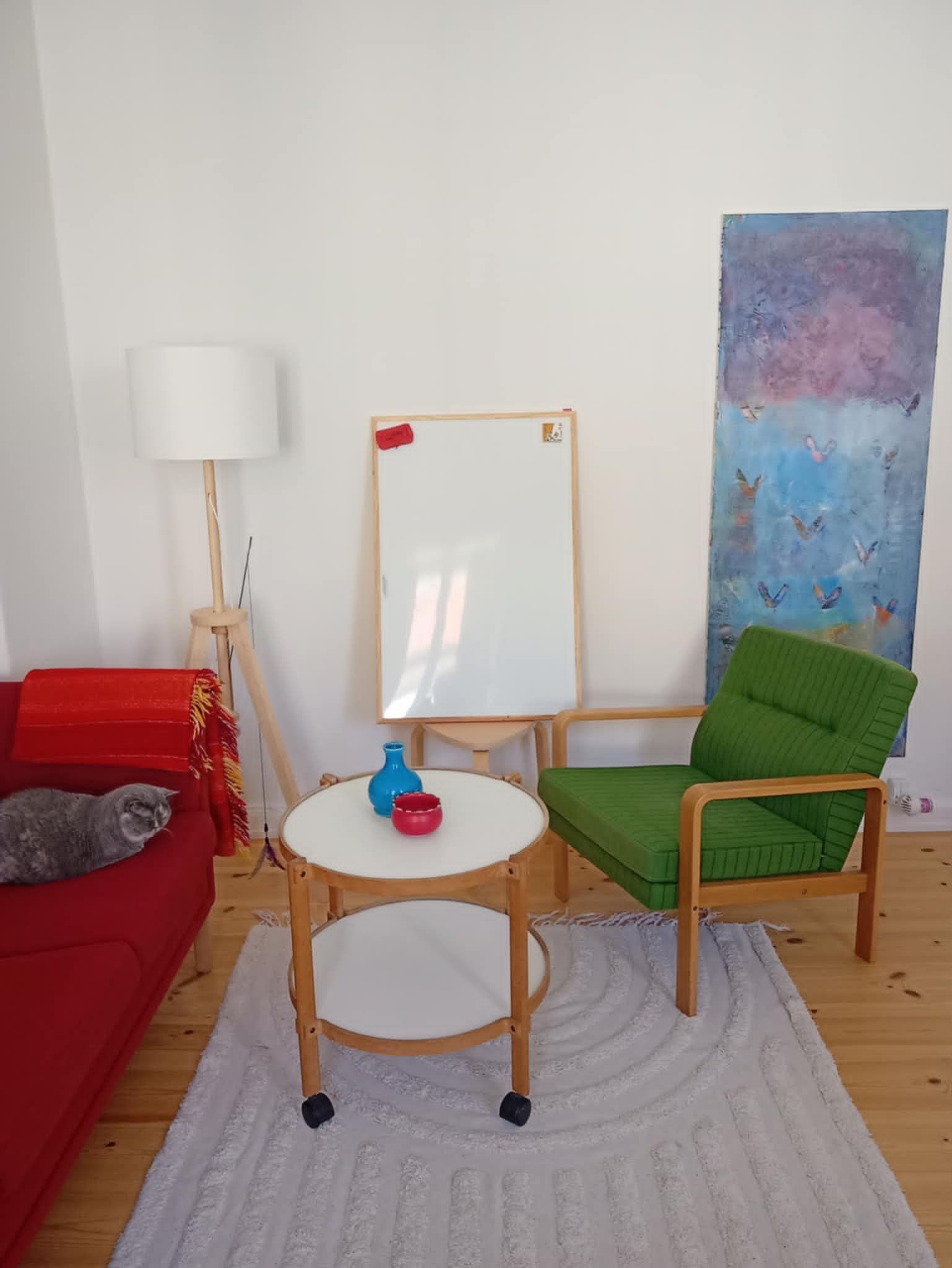 Et stuehjørne med en rød sofa med en grå kat liggende på, en hvid oval sofabord med blå og røde dekorative genstande, en grøn lænestol, en gulvlampe, et hvidt whiteboard, et vægkunstværk med flamingoer, en grøn puf og en hvid tæppe på trægulvet.