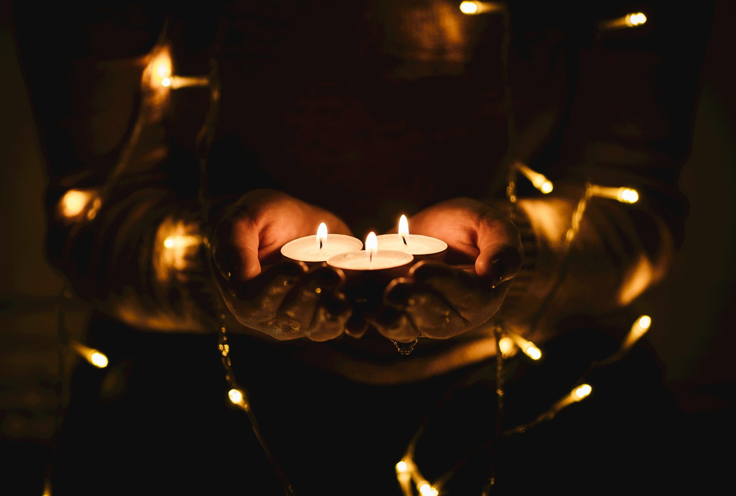 Person holding a tray with three lit candles, surrounded by string lights in a dark setting.