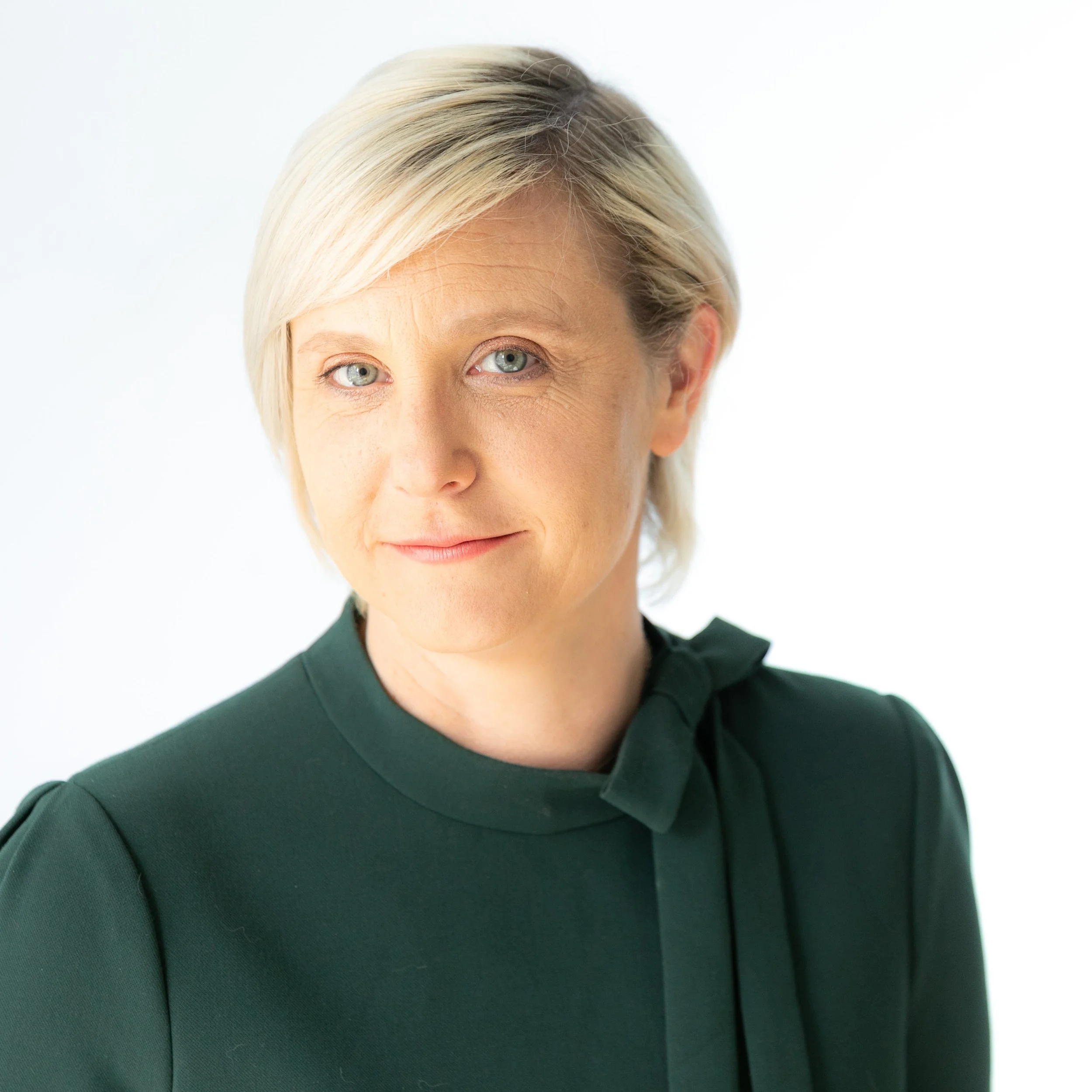 Close-up of a woman with short blonde hair and light blue eyes, wearing a dark green top, against a white background.