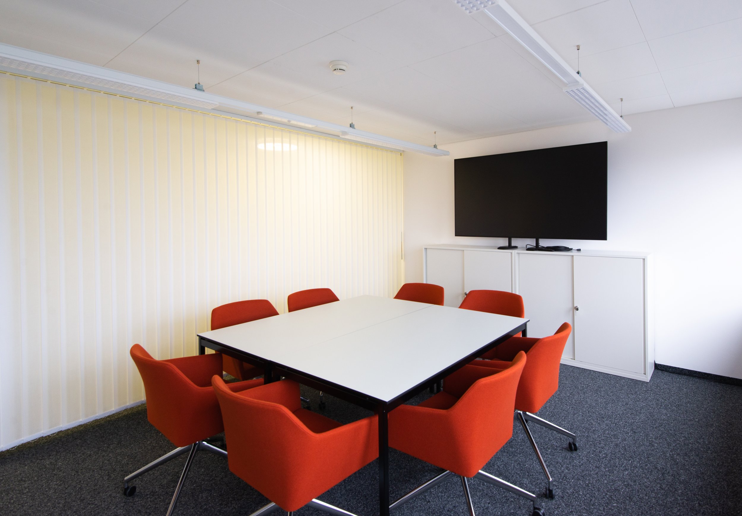 Ein Konferenzraum mit einem weißen Tisch, rot gepolsterte Stühle, einem schwarzen Flachbildschirm an der Wand, einer weißen Schrankeinheit und einer vertikalen, cremefarbenen Wand mit vertikalen Streifen.