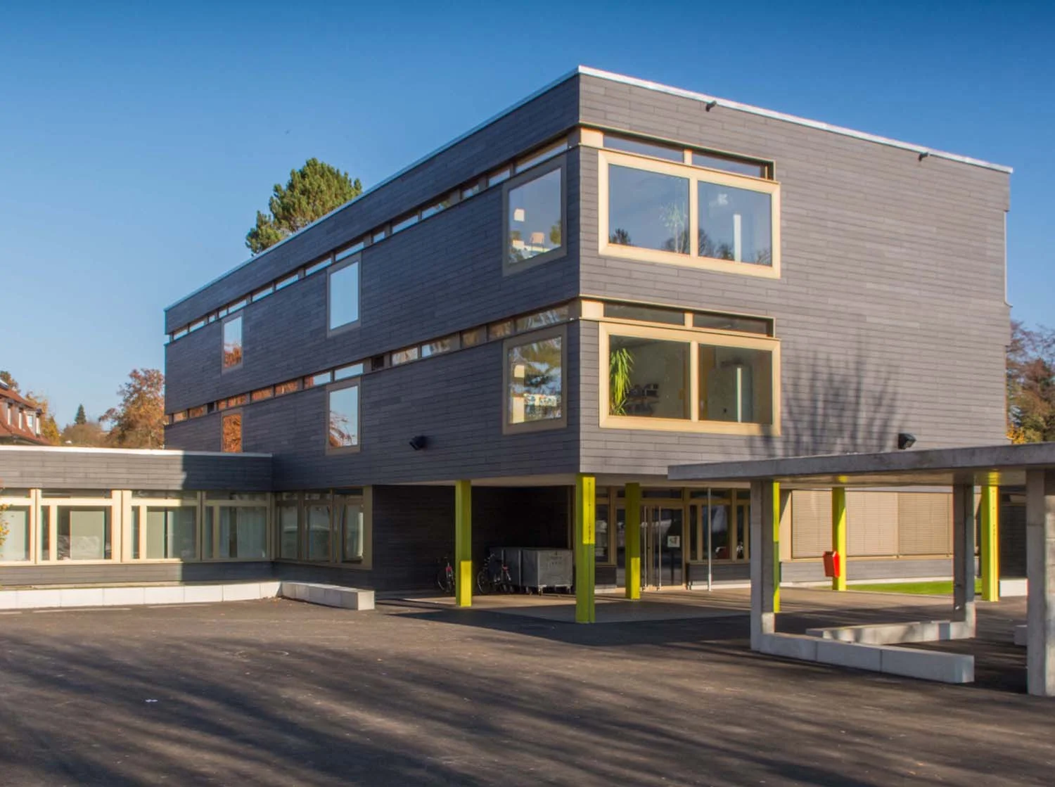 Modernes, mehrstöckiges Gebäude mit dunklem Holzsiding, großen Fenstern und bunten Stützpfeilern im Parkhausbereich, bei sonnigem Wetter.