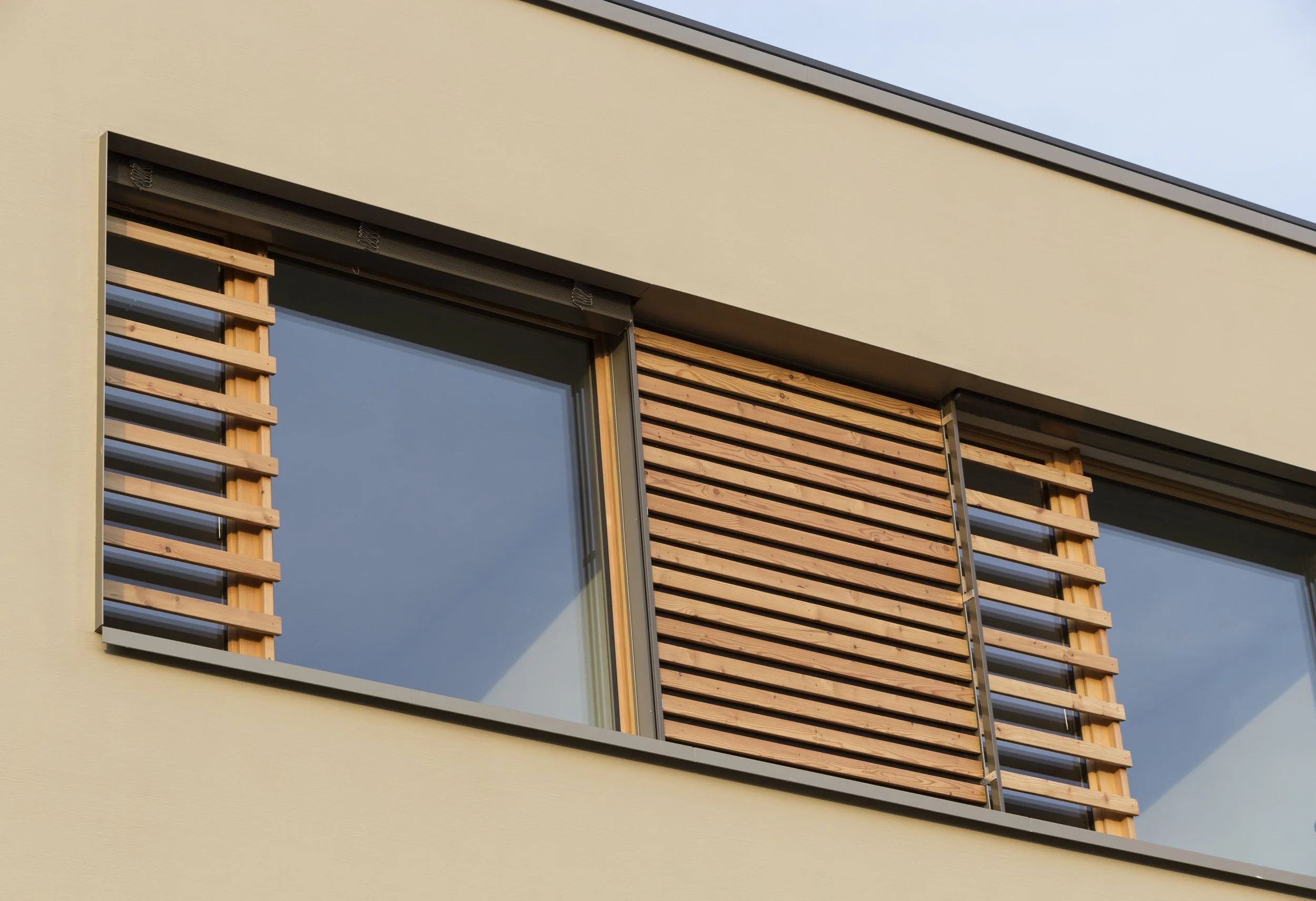Ansicht eines modernen Gebäudes mit großen Fenstern, Holzverkleidung und einer sandfarbenen Außenfassade, kurze Perspektive, klare Linien, Himmel im Hintergrund.