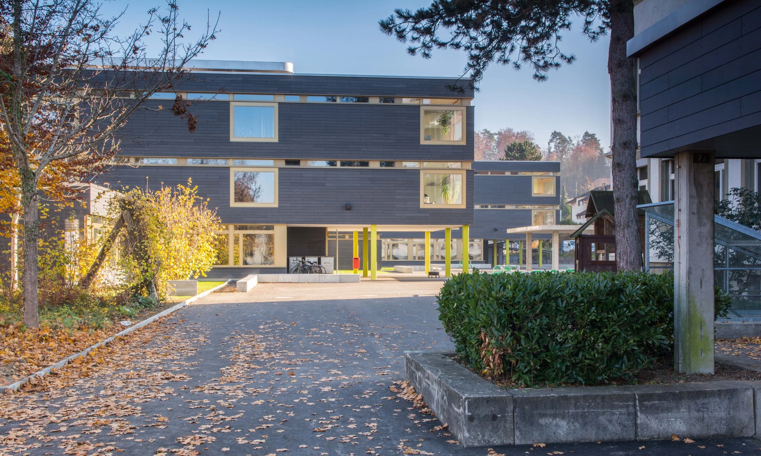 Ein moderner Gebäudekomplex mit mehreren Etagen, dunkelgraßen Fassaden, großen Fenstern und einem überdachten Eingangsbereich. Im Vordergrund befinden sich Bäume, herbstliche Blätter auf dem Boden und ein kleiner Fahrradständer.