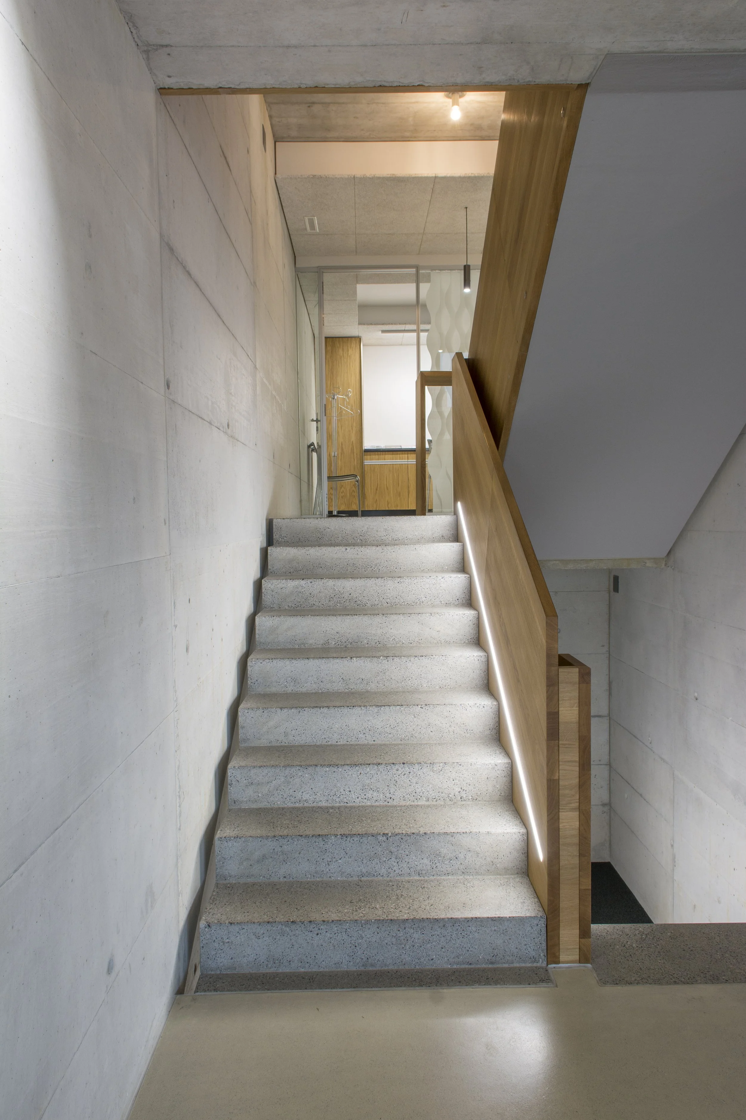 Ein moderner, minimalistisch gestalteter Innenraum mit einer Treppe, einer Holzgalerie und weißen Wänden, in einem Gebäude. Die Treppe ist aus Beton oder Stein, und die Galerie hat eine Holzbrüstung. Es gibt eine indirekte Beleuchtung unter der Holzgalerie.