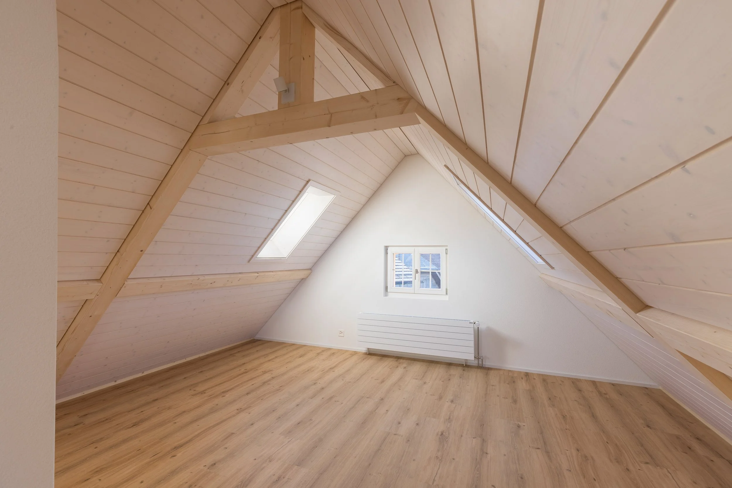 Ein heller Raum mit schwarzem Holzboden, weißem Heizkörper, zwei Dachfenstern und einer weißen Wand, mit sichtbaren Dachbalken aus Holz.
