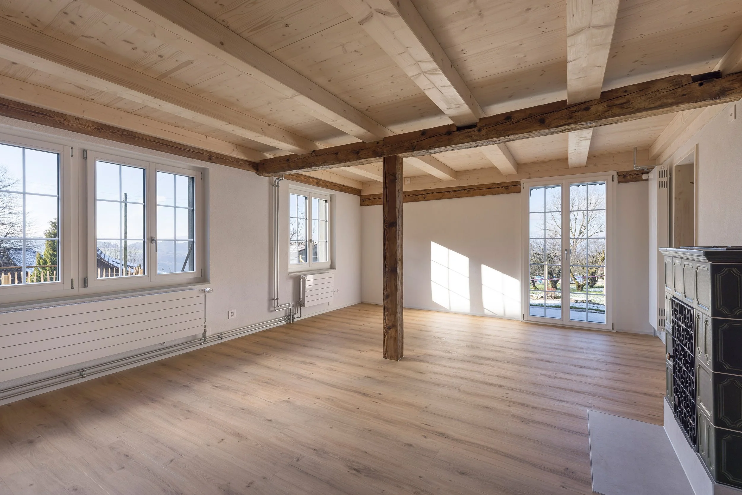 Innenraum eines hellen Zimmers mit Holzboden, weißen Wänden, großen Fenstern und sichtbaren Holzbalken an der Decke.