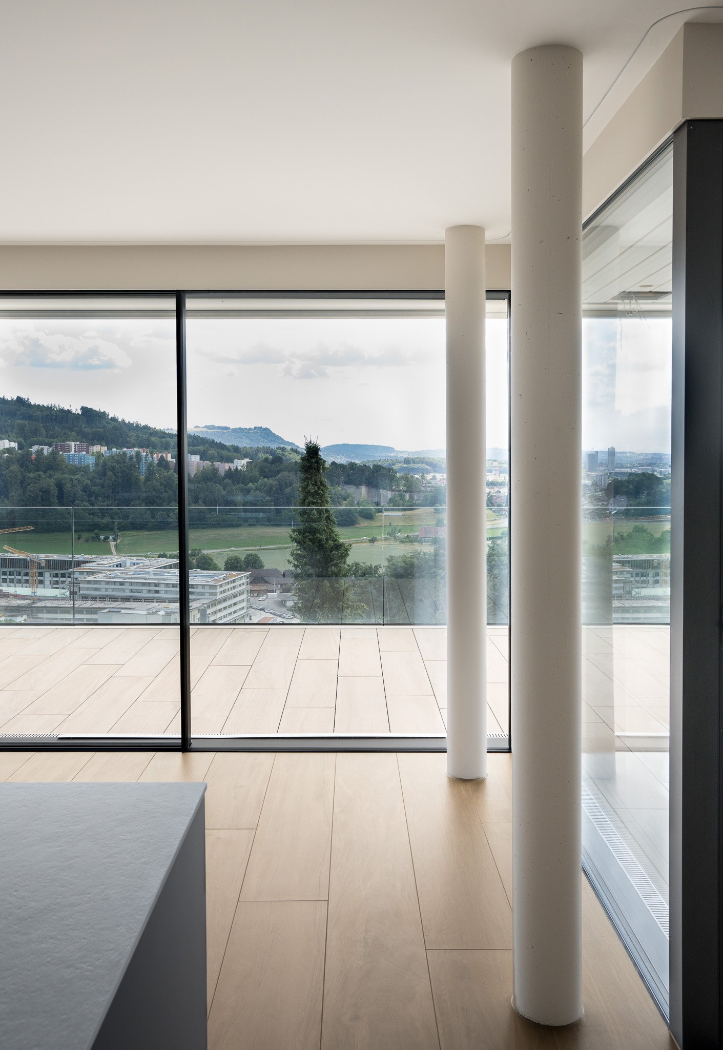 Innenraum eines modernen Gebäudes mit großen Fenstern, Blick auf eine Stadt und grüne Hügellandschaft, weiße Säulen, Holzboden und Minimaldesign.