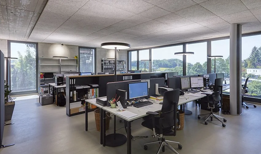 Offener Büroarbeitsbereich mit mehreren Schreibtischen, Computern, Stühlen und großen Fenstern, die eine grüne Landschaft zeigen.