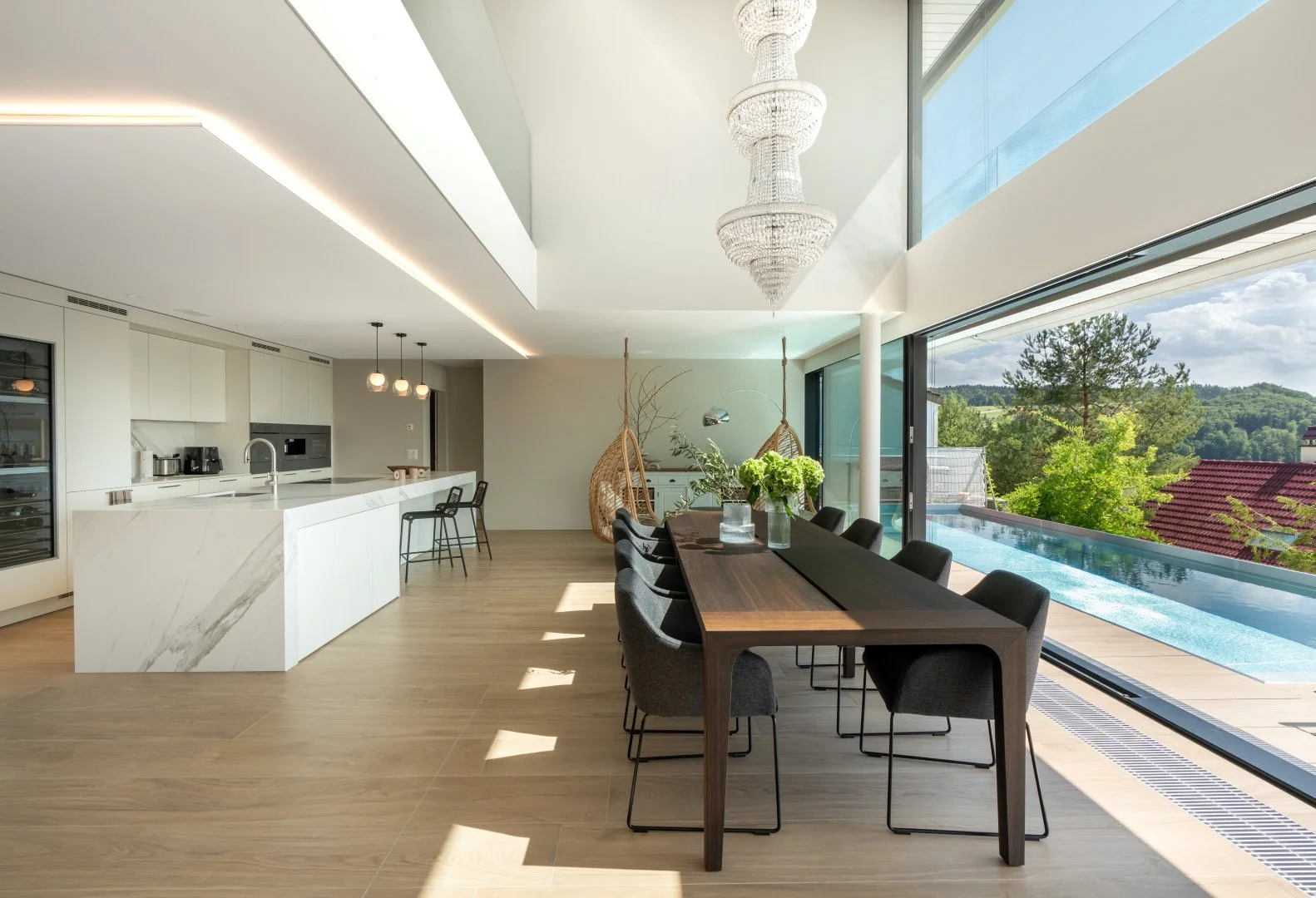 Moderne offene Küche und Essbereich mit Blick auf Natur im Hintergrund, große Glasfenster, heller Boden, weiße Wände, elegante Beleuchtung, Kissen und Pflanzen als Dekoration.