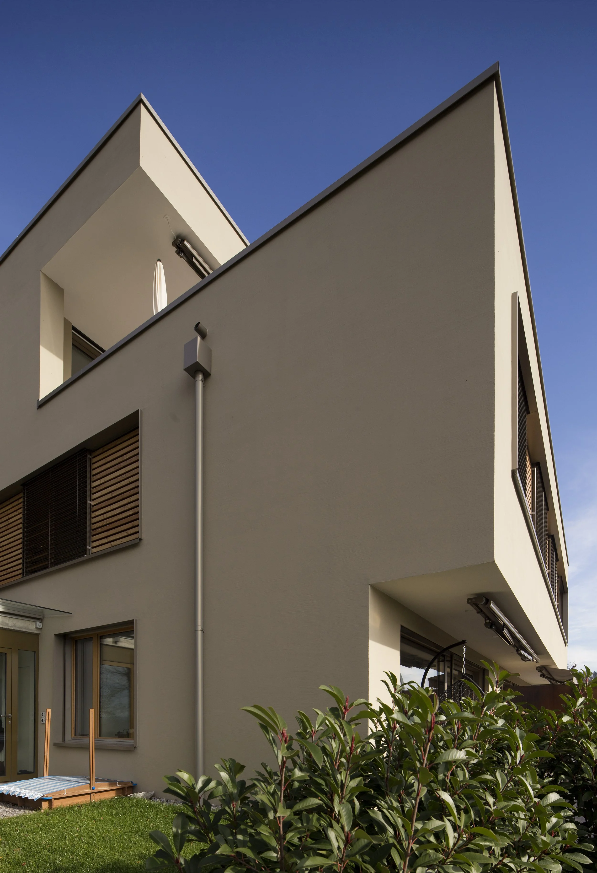 Ansicht eines modernen weißen Mehrfamilienhauses mit Balkon und Klappläden vor einem blauen Himmel, im Vordergrund Busch mit grünen Blättern.