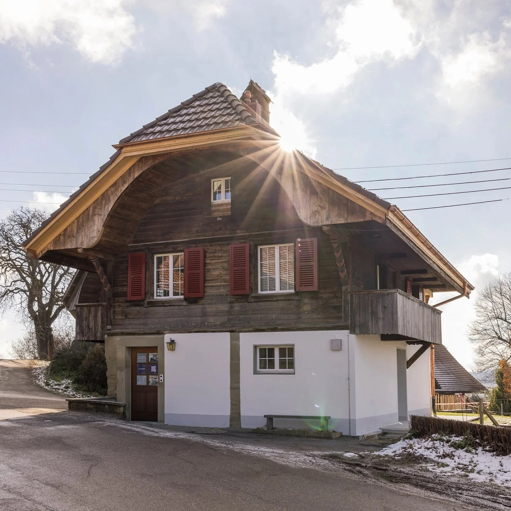 Haus mit Holzfassade, roten Fensterläden und einem weißen Sandstein-Giebel, unterhalb eines Daches mit Schindeln. Die Sonne scheint hinter dem Dach, während Wolken den Himmel bedecken.
