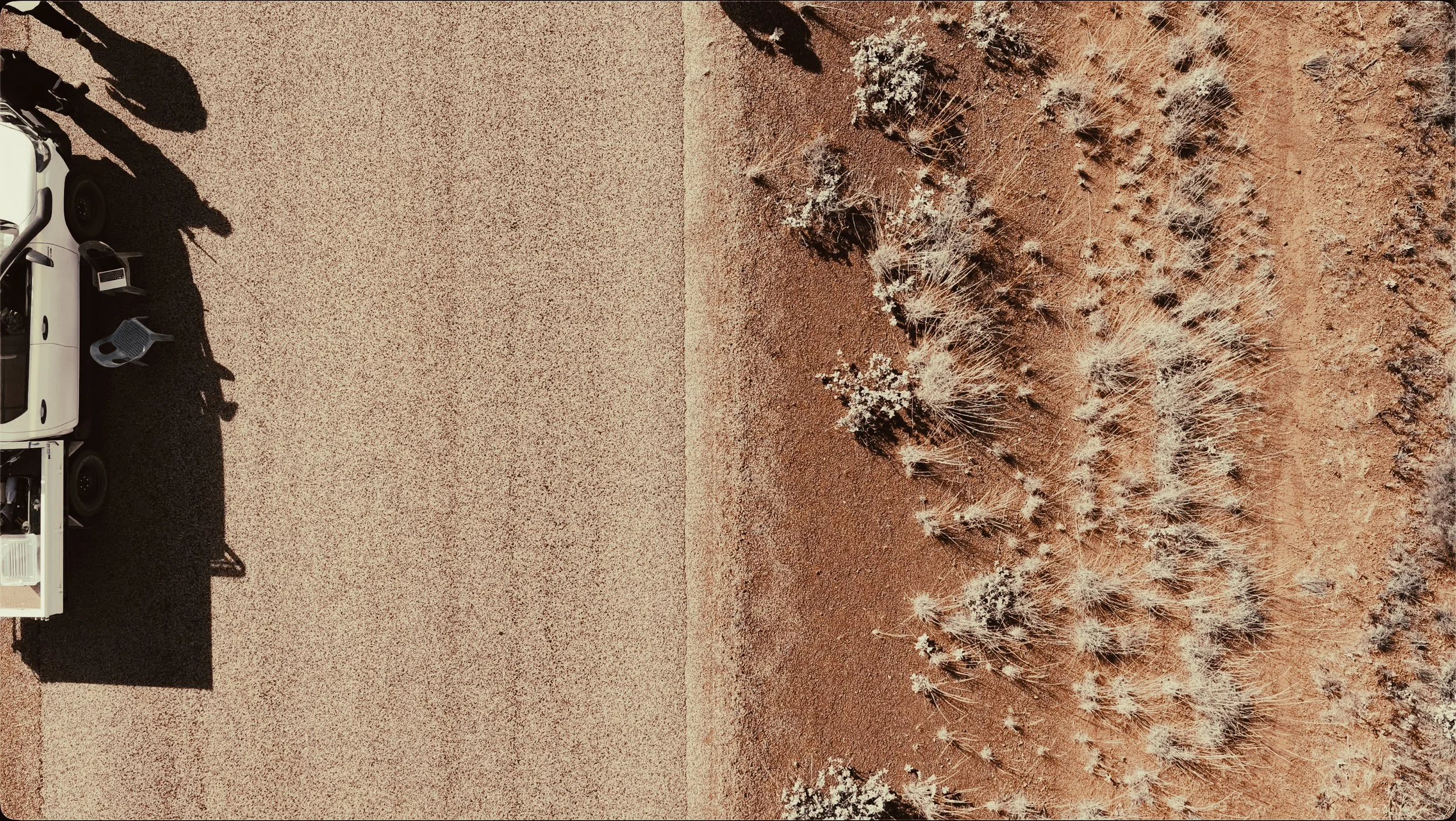 Side view of a white vehicle parked on a gravel surface with desert vegetation and dry bushes nearby.