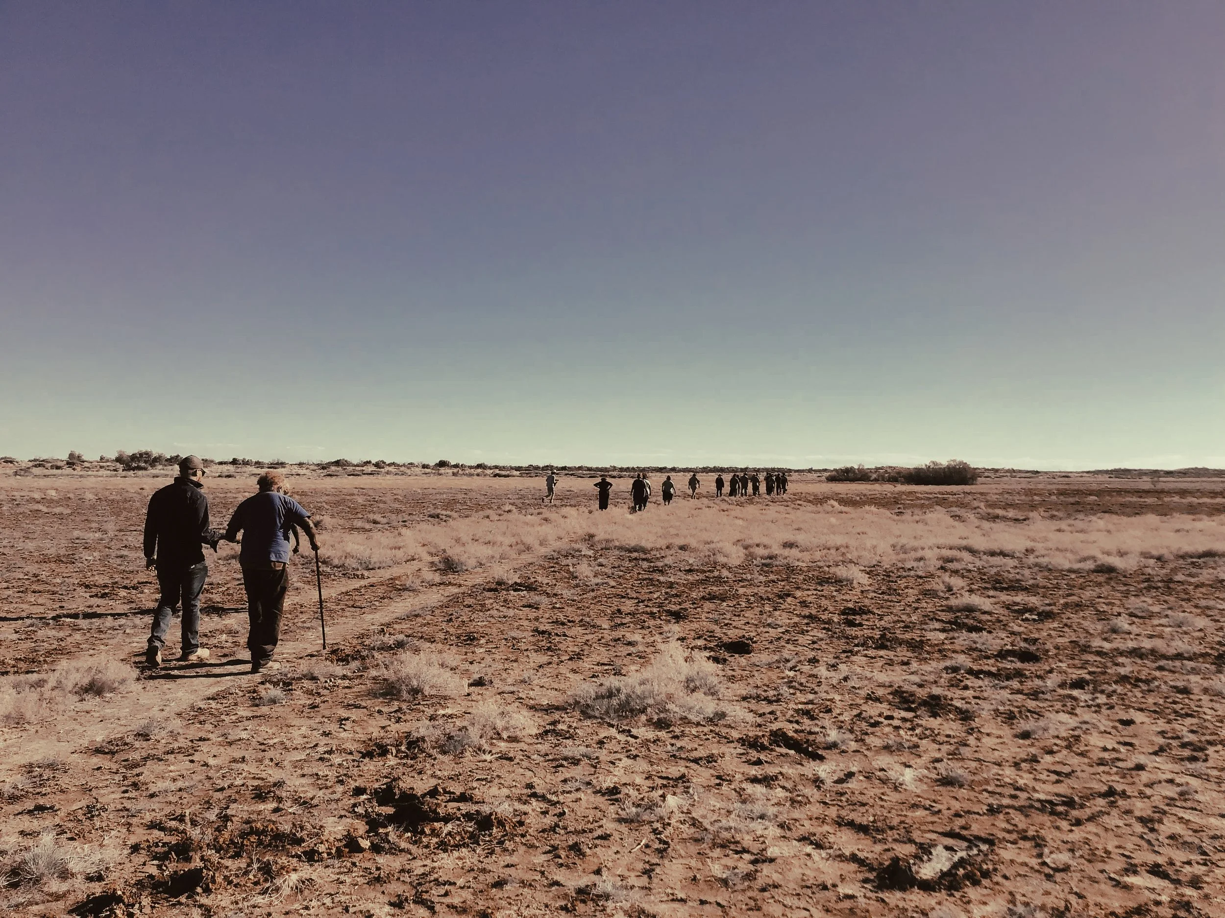 A group of people walking across a dry, desert landscape under a vast, clear sky.