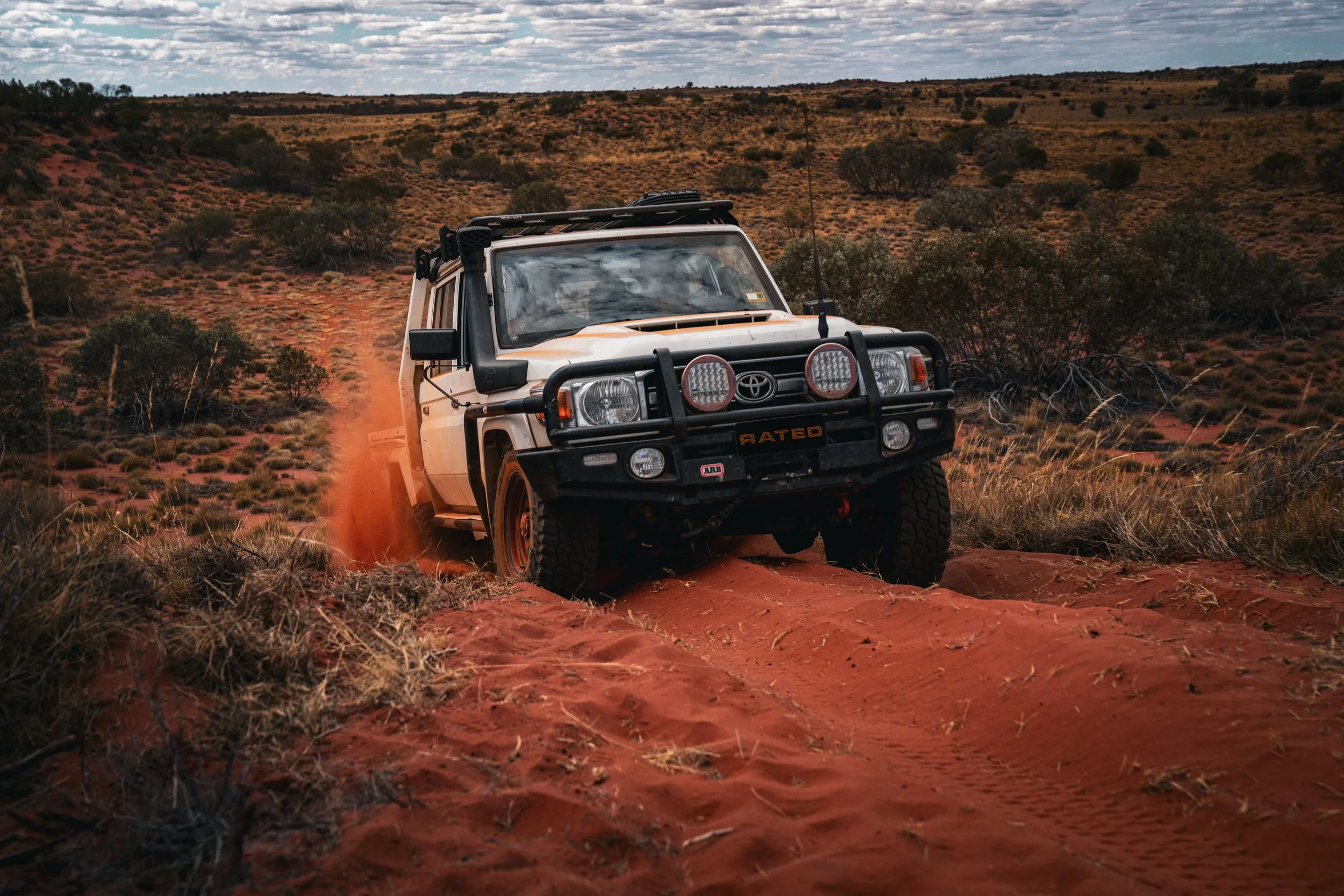 A white Toyota off-road vehicle driving through a desert landscape with reddish sand and sparse vegetation under a partly cloudy sky.