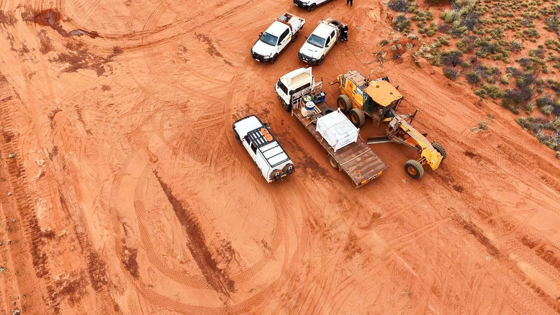 Aerial view of a construction site with multiple white pickup trucks, a yellow construction vehicle, and other equipment on reddish dirt, with sparse green vegetation on the right side.