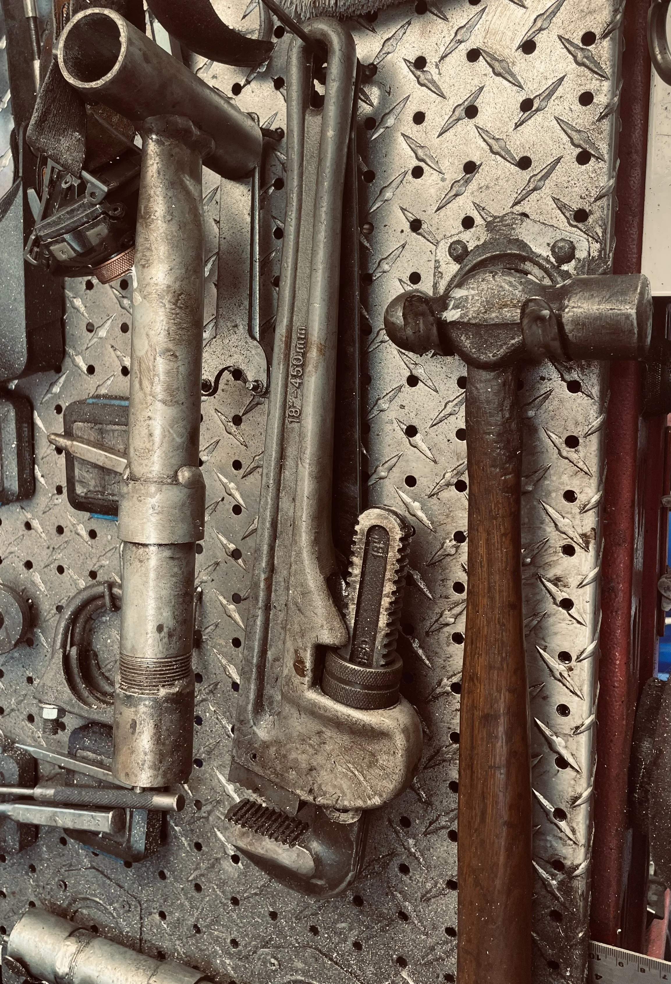 A collection of industrial tools is laid out on a metal diamond plate surface. The tools include a large metal pipe, a metal rod, a hammer with a wooden handle, and various smaller tools and parts. The surface has a pattern of raised diamonds and is within a workshop or garage setting.