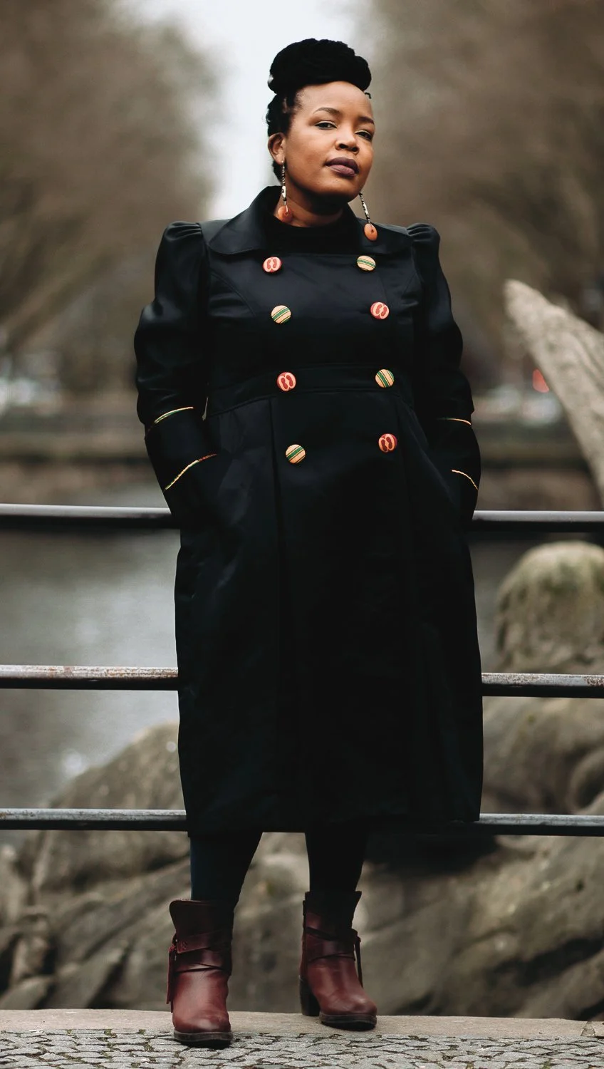 A woman standing outdoors on a bridge with a railing, wearing a black coat with embellished buttons, burgundy ankle boots, and long earrings, with a blurred background of trees and water.