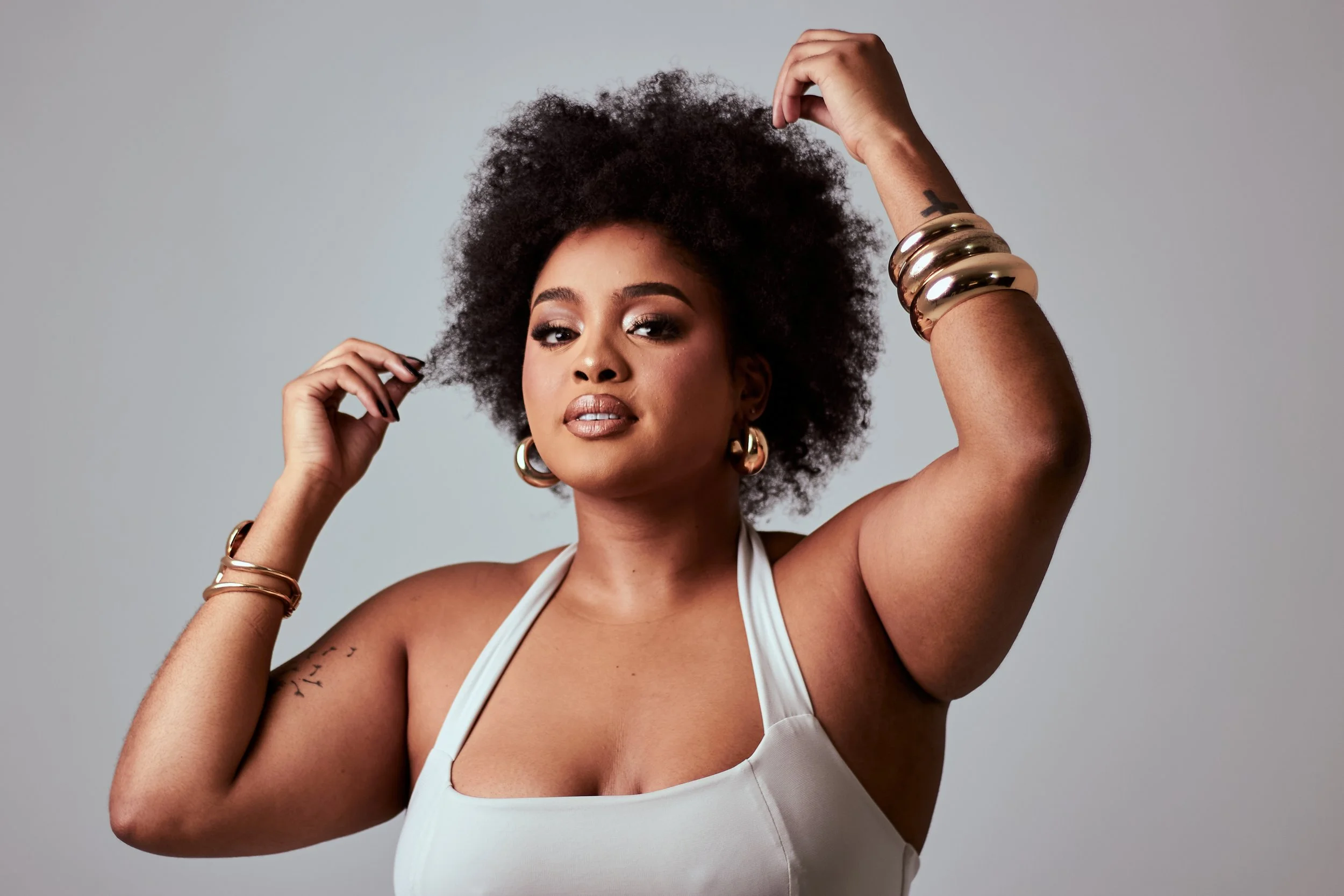 A woman with dark skin and natural curly hair posed against a plain background, wearing gold jewelry including large hoop earrings and multiple bangles, and a light-colored top with thin straps.