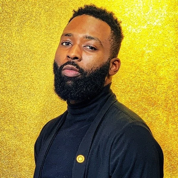 A young Black man with a beard and short curly hair, wearing a black turtleneck and a black jacket, standing against a shiny gold background.
