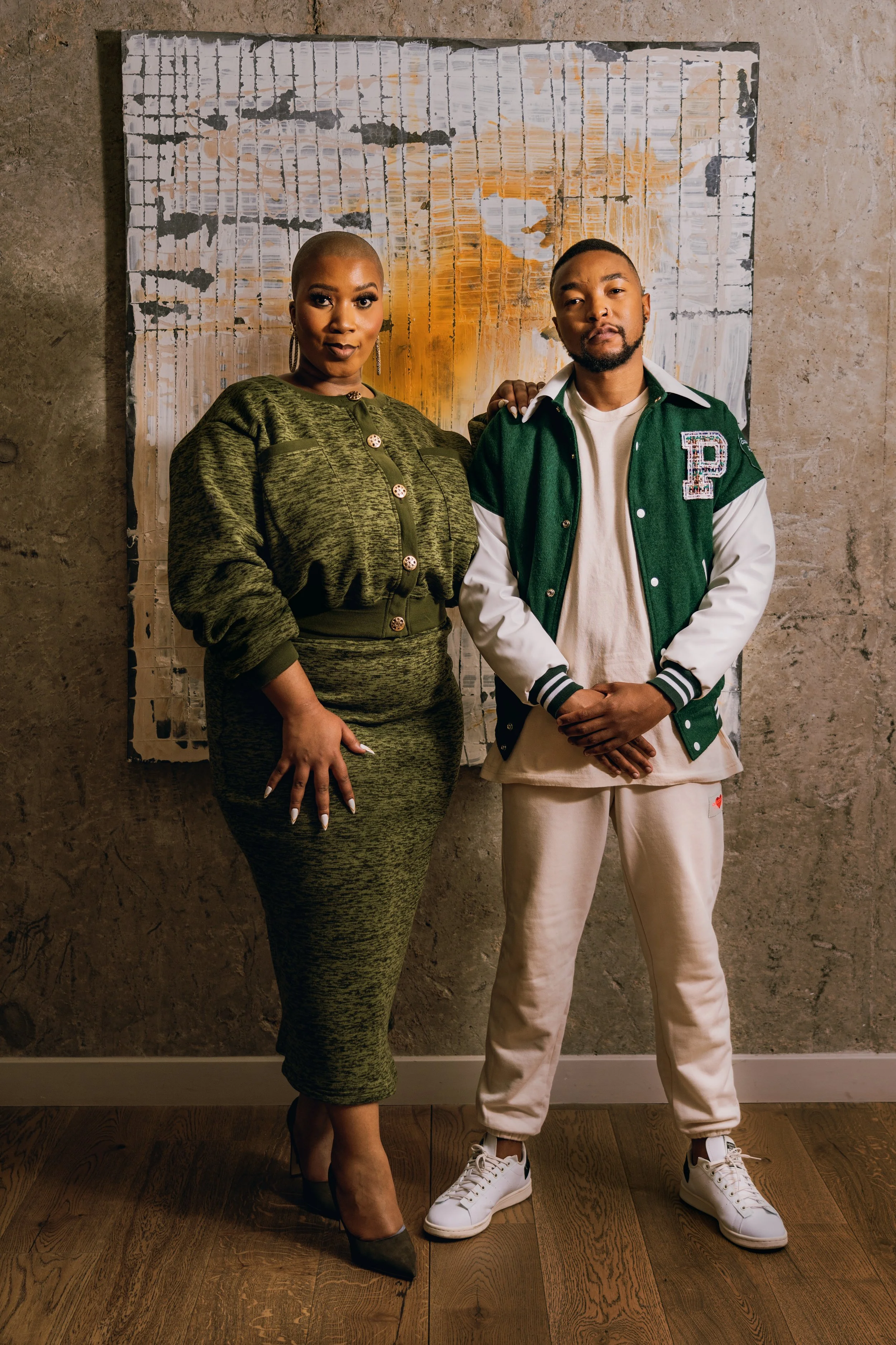 Two people standing in front of a textured artwork on a concrete wall. The woman on the left is dressed in a green patterned dress with buttons, wearing high heels and hoop earrings. The man on the right is dressed in a green and white varsity jacket, white t-shirt, cream sweatpants, and white sneakers. He has a beard and short hair.