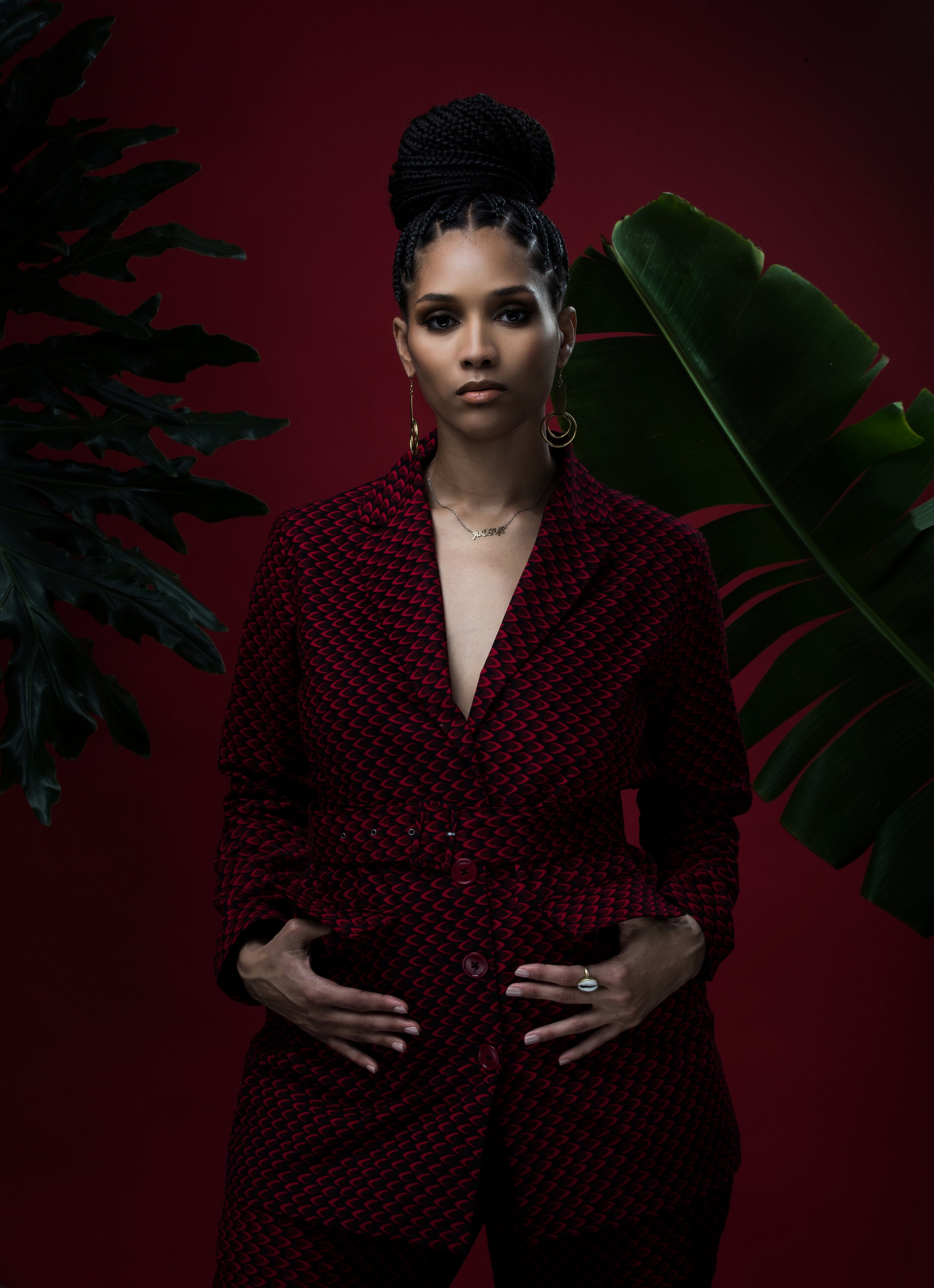 A woman with dark braided hair styled in an updo, wearing a red and black patterned suit, gold hoop earrings, a necklace with the word 'LOVE,' and a ring, standing against a deep red background surrounded by large green tropical leaves.