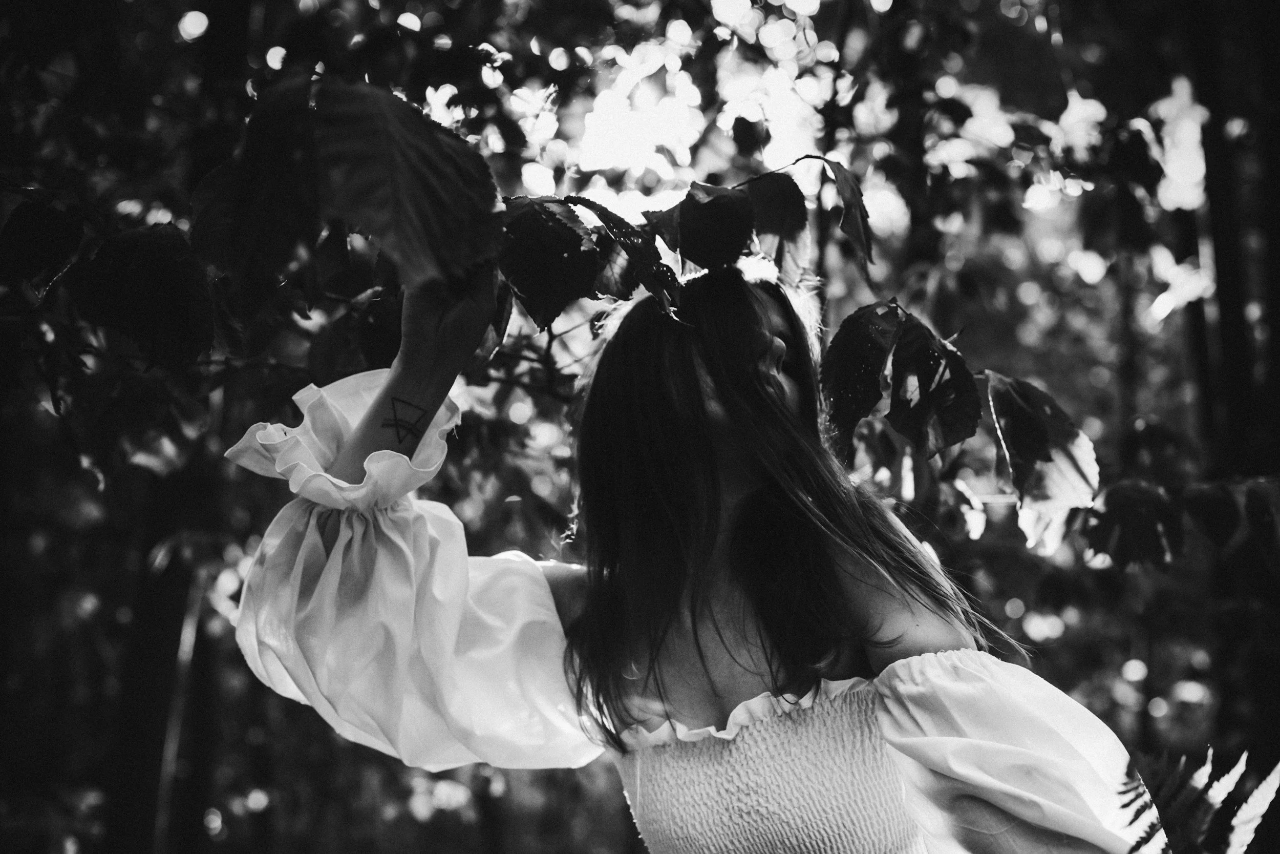 A child in a long dress reaches up toward branches of a tree with leaves, backlit by sunlight, in a black-and-white photo.