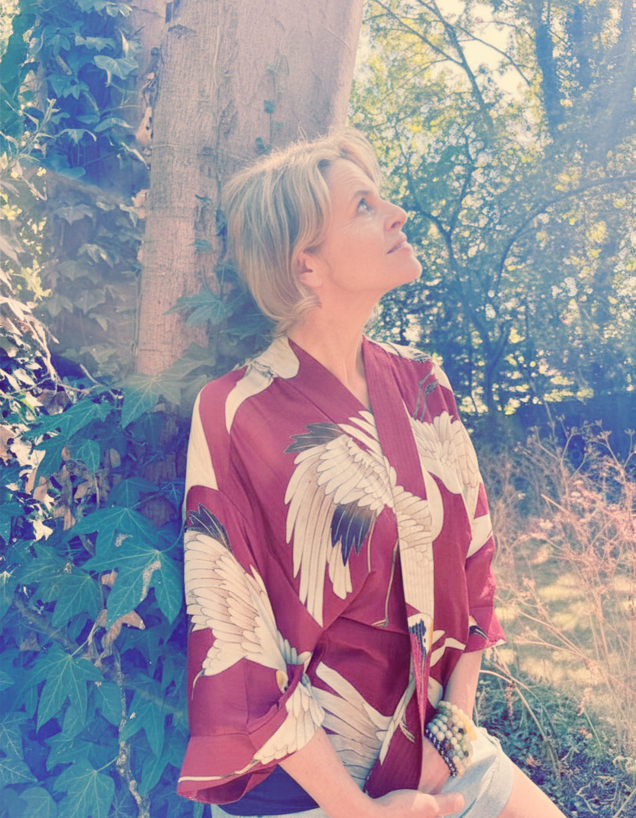 A woman with blonde hair wearing a floral kimono jacket sitting outdoors next to a large tree, with sunlight filtering through the leaves.