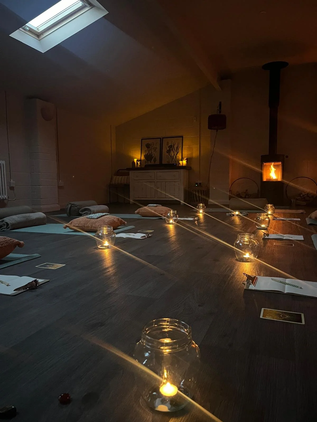 A cozy indoor space set up for a meditation or yoga session, with cushions, towels, and candles arranged on a wooden floor, a warm fireplace on the right, and a skylight providing natural light, creating a tranquil atmosphere.