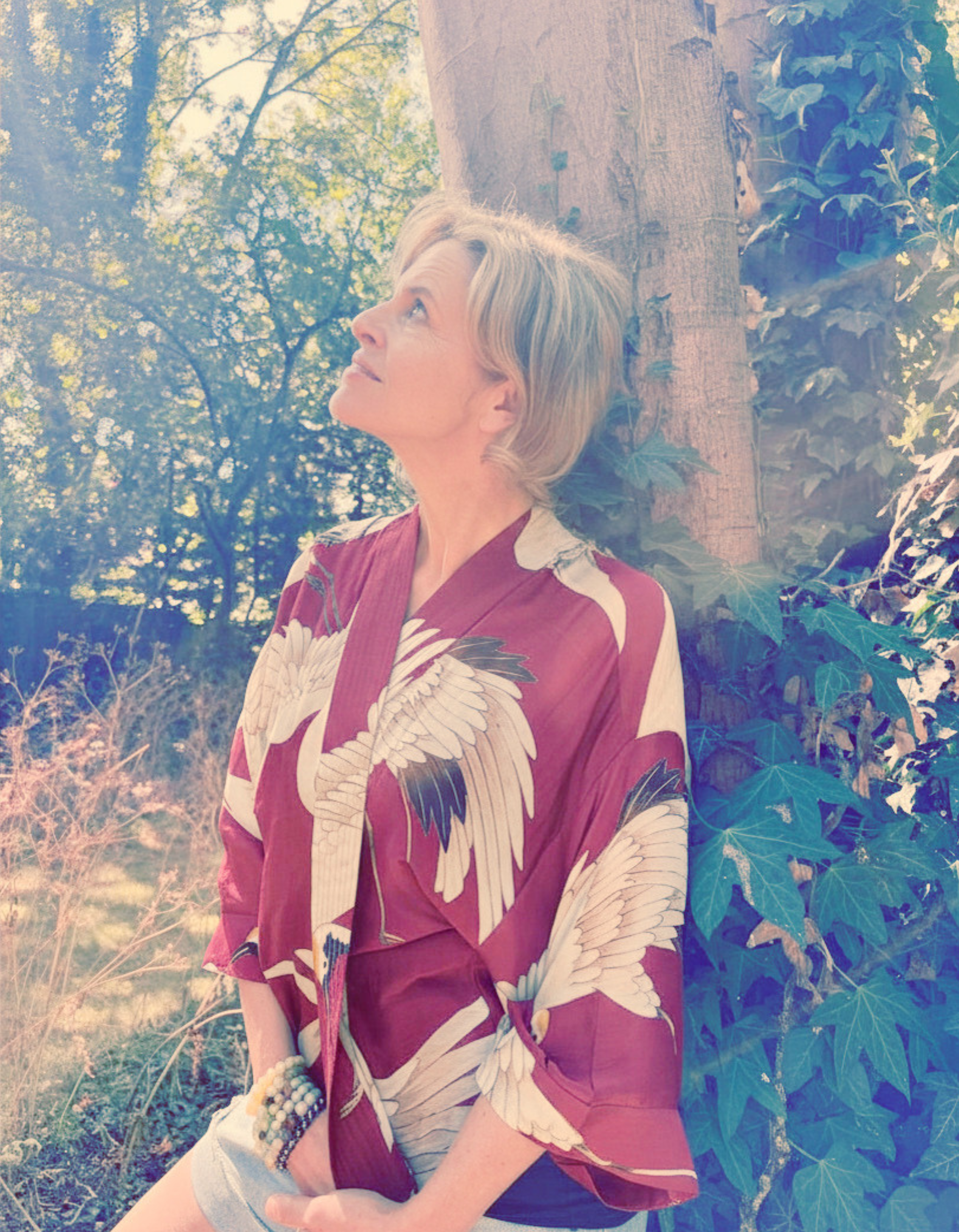 A woman with short blonde hair leaning against a tree in a sunny outdoor setting, wearing a red kimono-style top with a white crane pattern, surrounded by greenery and ivy.