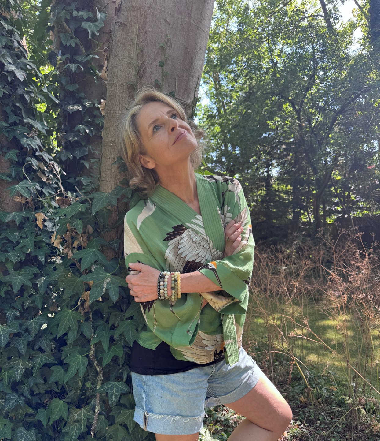 A woman with blonde hair in a green kimono-style top with bird illustrations, standing outdoors near a tree with ivy, crossing her arms and looking upwards with a thoughtful expression.