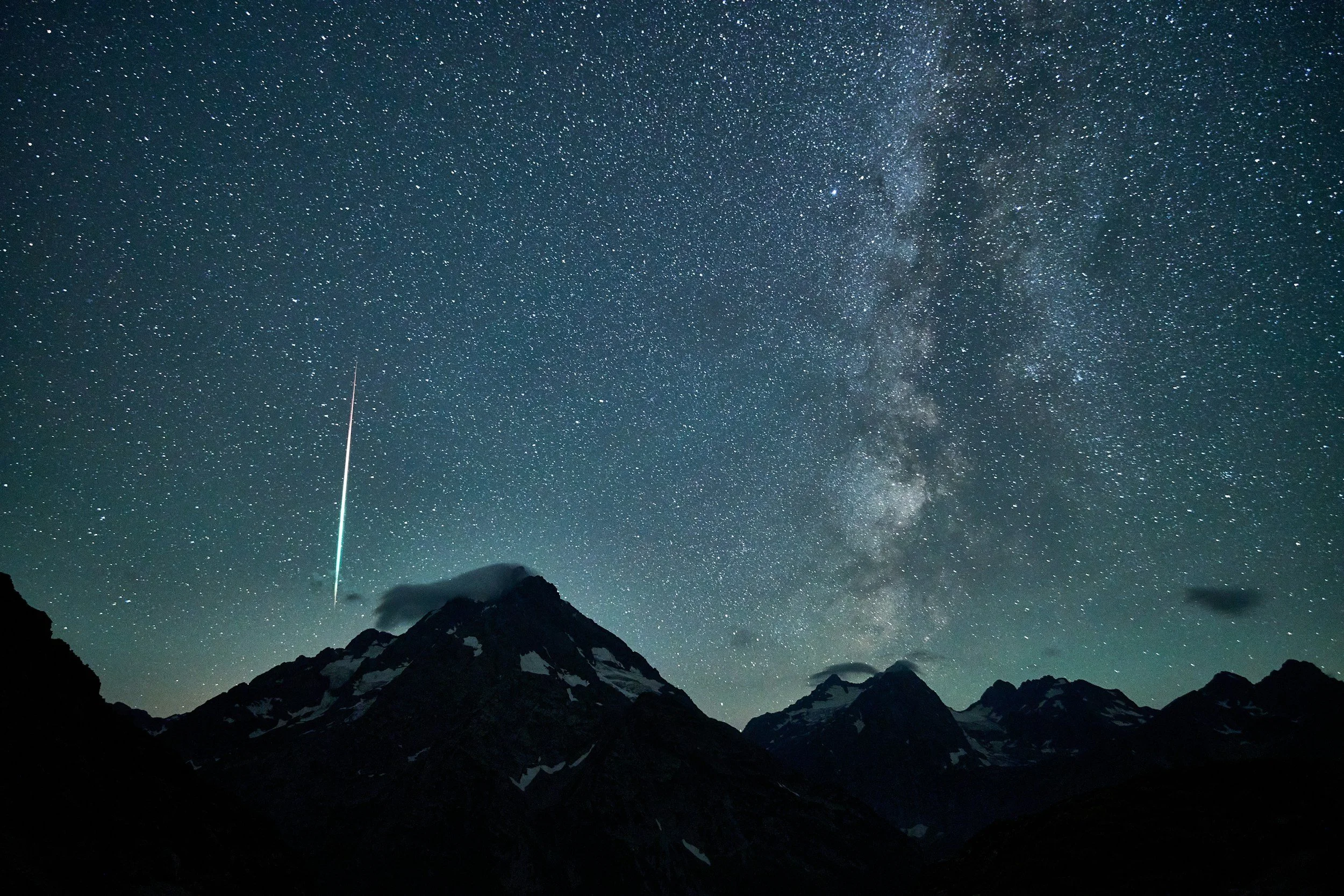 Night sky filled with stars over mountain range, with the Milky Way galaxy visible and a bright meteor streaking downward.