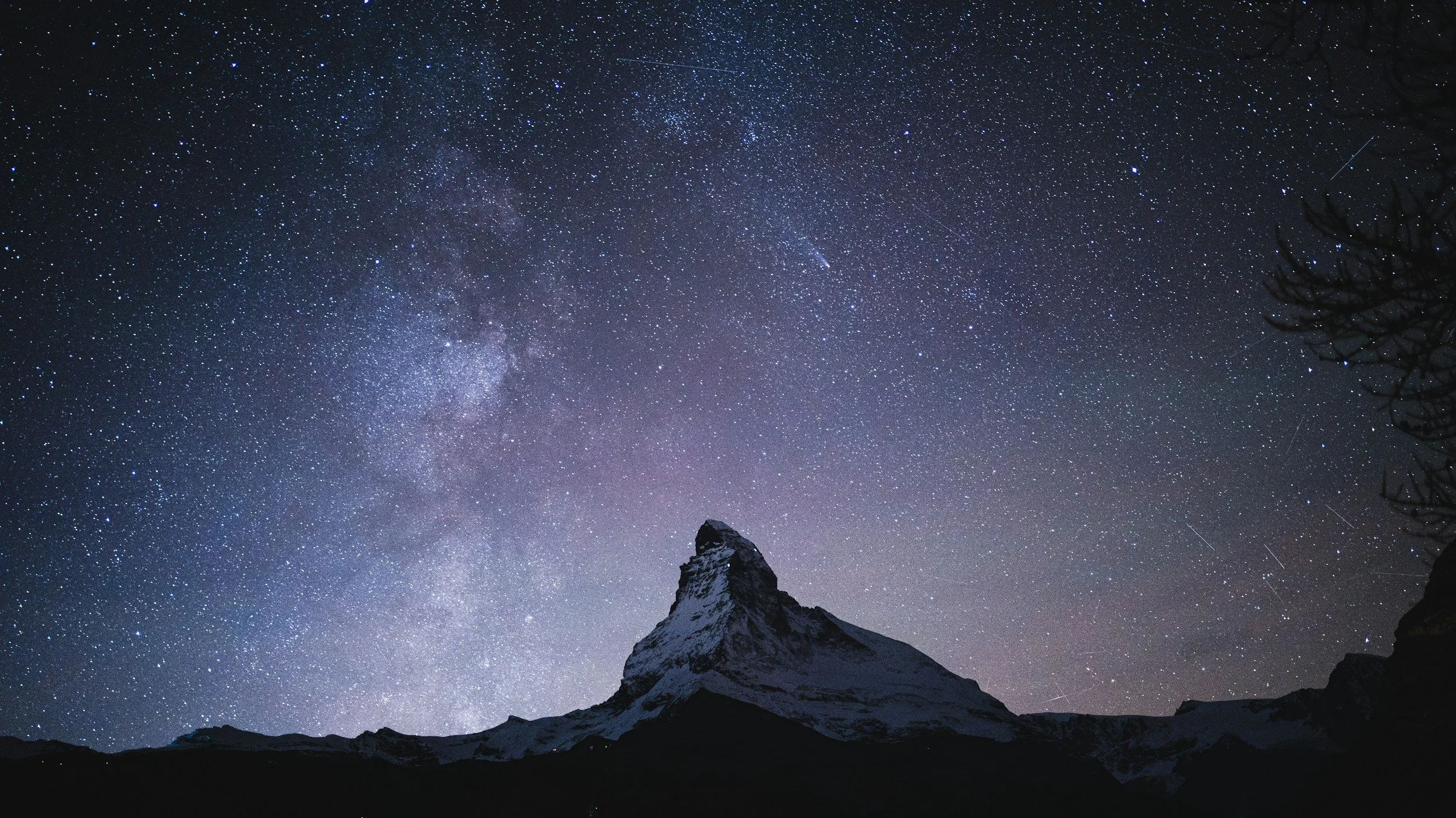 Starry night sky with numerous visible stars and a faint Milky Way galaxy over a snow-capped mountain peak.