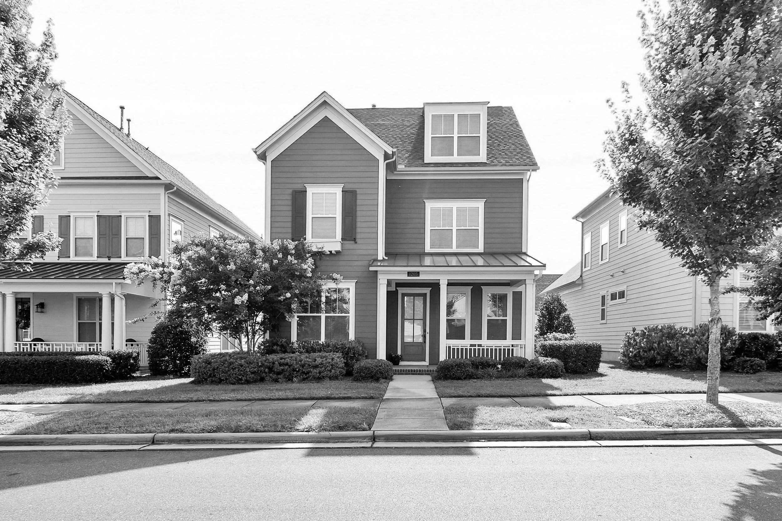 Black and white photo of a two-story house with a front porch, surrounded by grass, bushes, and trees, located on a street.