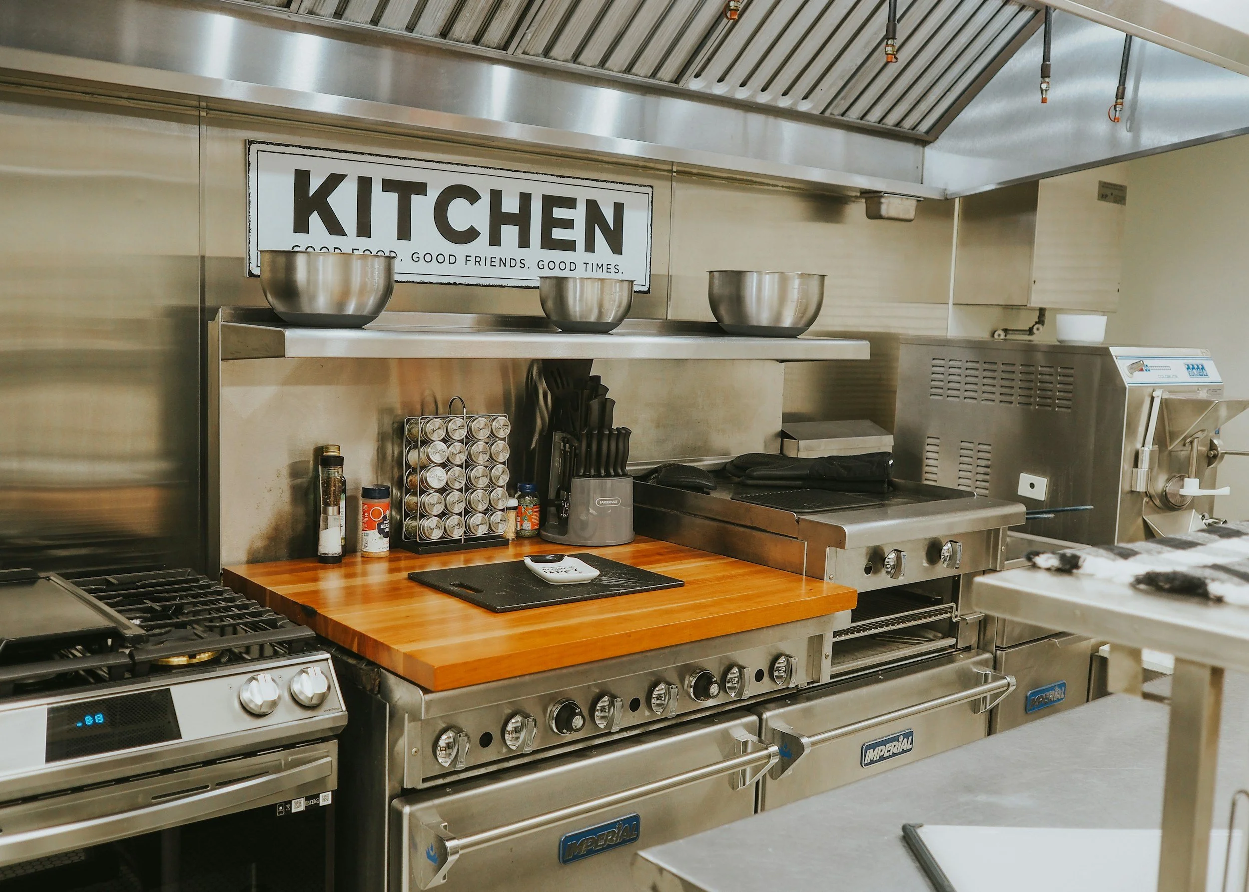 Commercial kitchen with stainless steel appliances, a wooden countertop, a spice rack, and a sign that reads ‘KITCHEN, GOOD FOOD, GOOD FRIENDS, GOOD TIMES’.