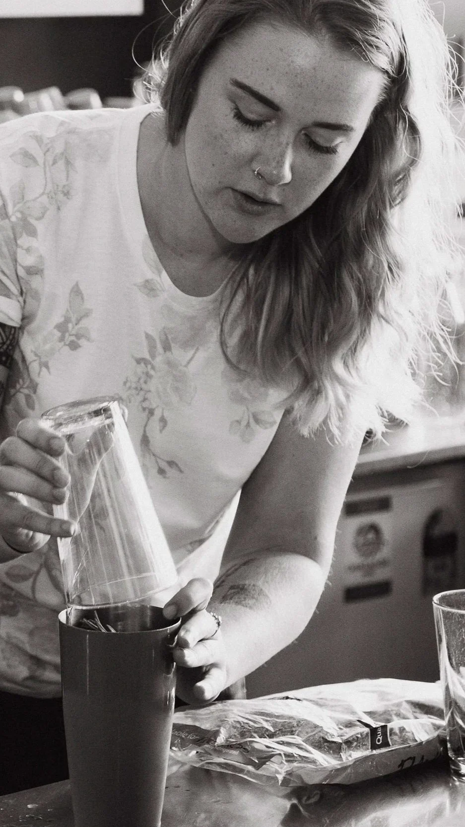 A woman with long wavy hair, freckles, and a nose ring is pouring from a glass into a tall mug at a table, with a plastic bag and a glass cup nearby.