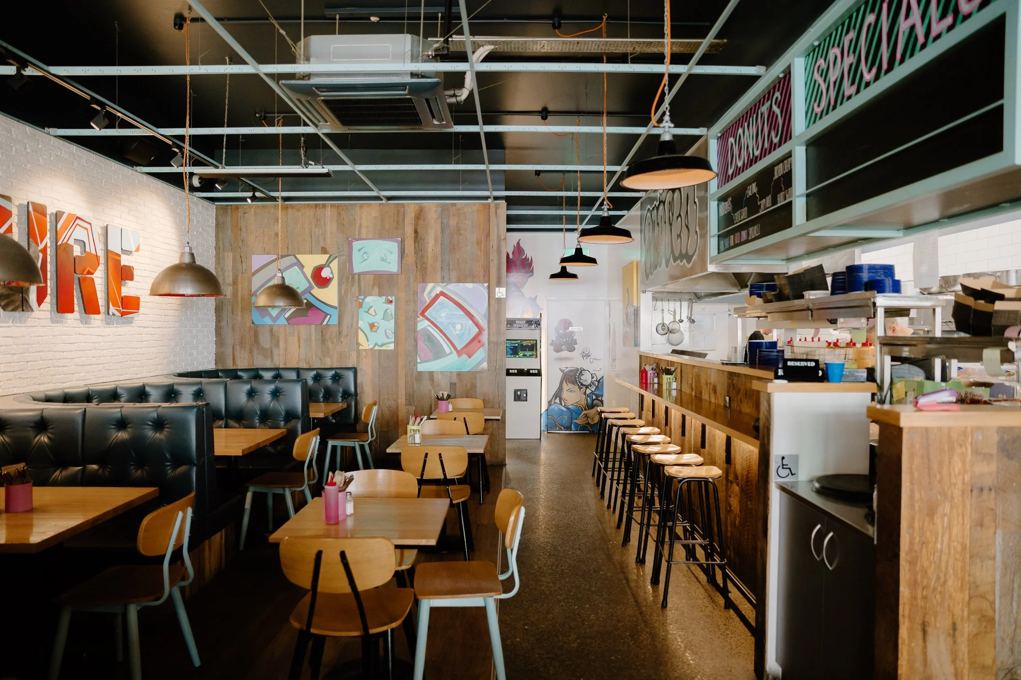 Interior of a modern casual restaurant with wooden tables, chairs, and black leather booth seating. Artworks and paintings decorate the white brick walls, with pendant lights hanging from the ceiling. The counter area with shelves displaying dishes and utensils is visible on the right side.